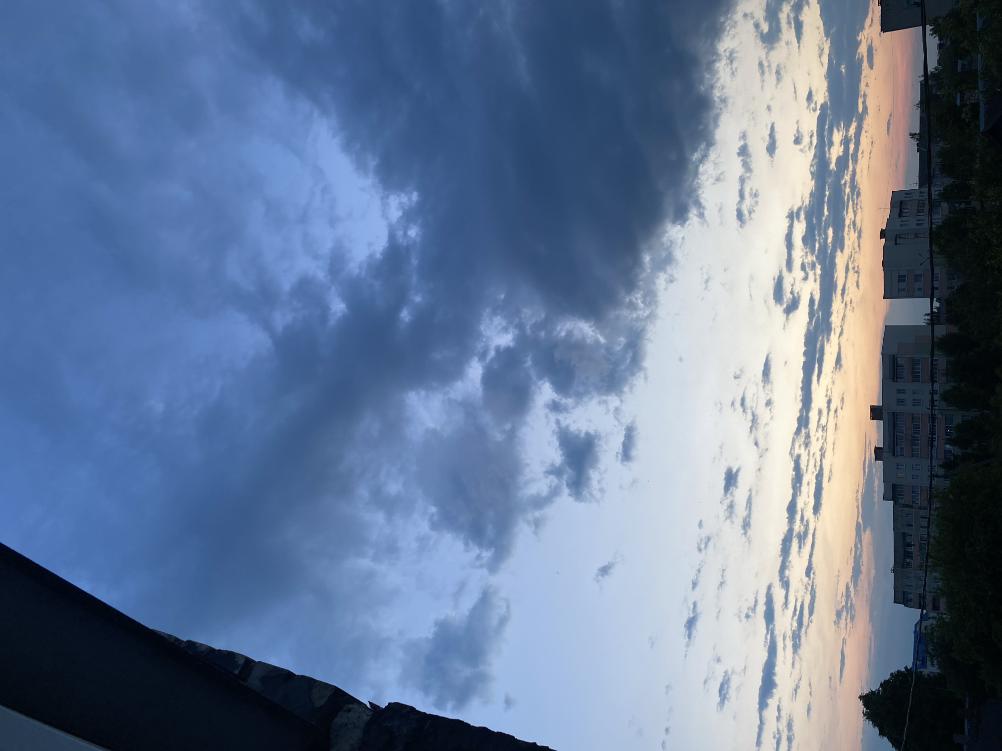 The image captures a view of the sky over the city of Krupki, Belarus, from what appears to be an elevated vantage point. The scene is dominated by a dramatic sky filled with clouds, transitioning from a deep blue and gray at the top to lighter shades of blue, pink, and orange towards the horizon, suggesting either dawn or dusk. In the lower portion of the image, several multi-story residential buildings are visible, characterized by their neutral facades and numerous windows. Silhouetted against the sky and the buildings are dense green trees. On the left side of the frame, a dark, angular structure, possibly part of a building or balcony, occupies a significant portion of the foreground, creating a framed view of the sky and city. No people or active events are depicted in the scene.