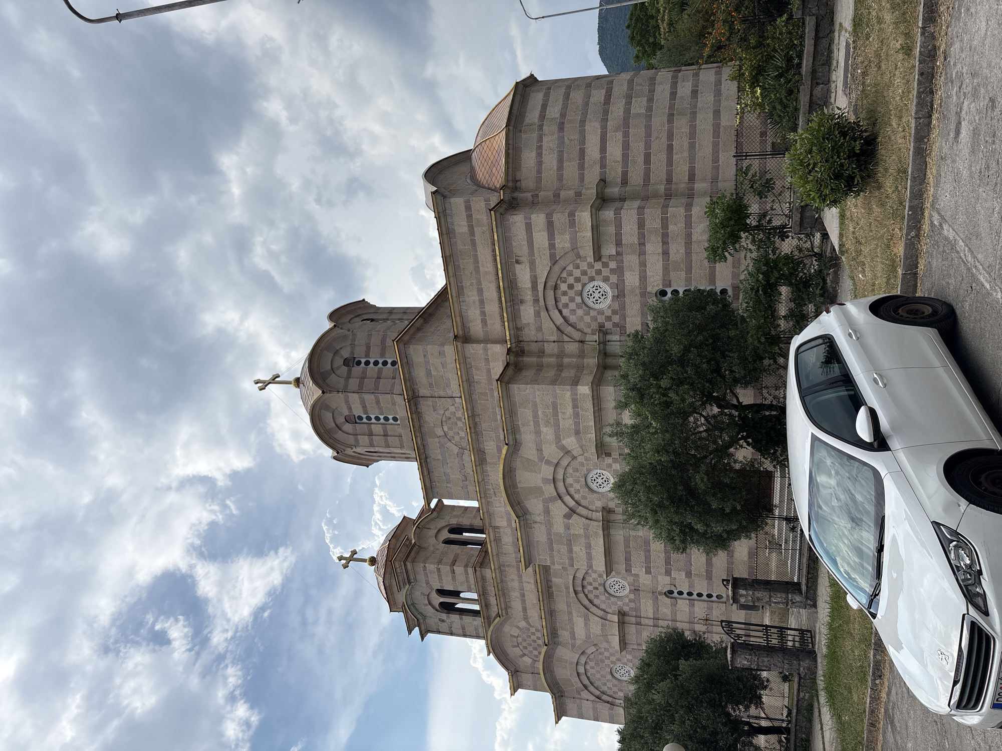 The image shows the exterior of a large stone church with striped patterns and domes. A white car is parked on the right side of the image, partially obscuring the lower portion of the church. The sky is cloudy, with patches of blue sky visible. Trees and greenery surround the church. The time of day appears to be daytime. There is no visible text in the image.