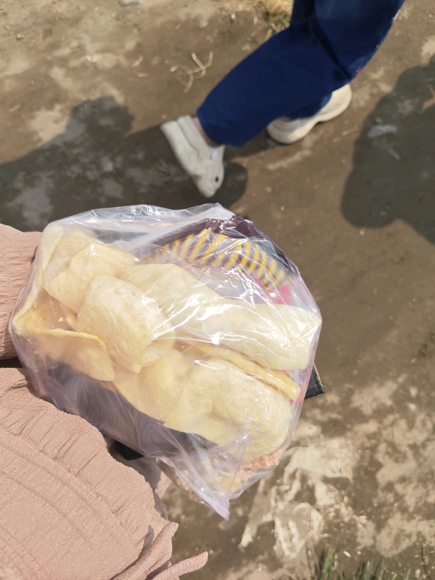 In Bangsal, Indonesia, a clear plastic bag filled with light-colored, puffy snacks, possibly crackers or chips, is held by a person wearing a tan-colored textured fabric garment. The bag is translucent, revealing the food items inside, some of which are stacked. Behind the bag and the person's hand, a second person's legs are visible, clad in dark blue trousers and white sneakers. Their feet are casting shadows on the dusty, mottled ground. The ground appears to be outdoors, possibly a pathway or a dirt area, with scattered debris. The scene is illuminated by natural light, suggesting it is daytime.