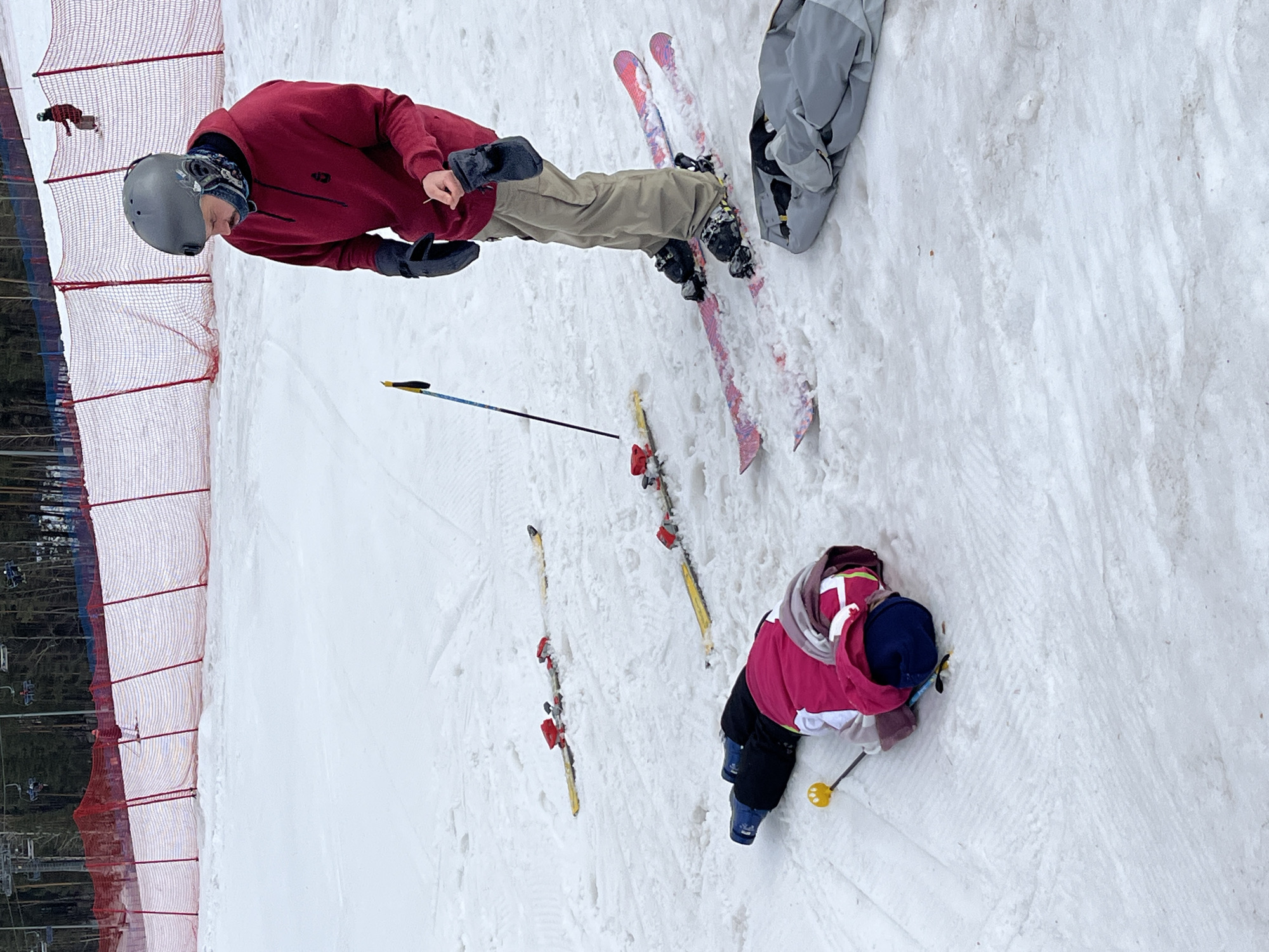 The scene depicts a snow-covered ski slope at an unidentified ski resort, location Unknown, Unknown. In the foreground, an individual wearing a grey helmet, red hooded sweatshirt, khaki pants, and dark gloves stands upright on pink-patterned skis, looking downwards. This individual holds a small, light-colored object in their left gloved hand. Adjacent, a second individual, dressed in a pink jacket, black pants, and blue ski boots, lies prone, face-down on the snow. Two yellow skis with red bindings are detached from the fallen individual's boots and rest nearby in the snow. A single yellow-tipped ski pole is positioned near the prone person. Two additional ski poles are stuck vertically in the snow to the left of the standing individual. A grey soft bag rests on the snow to the right. The mid-ground features a red mesh safety net spanning the width of the slope. Behind the netting, a coniferous forest and ski lift infrastructure are visible. A third individual on skis or a snowboard is present in the distant background near the safety netting.