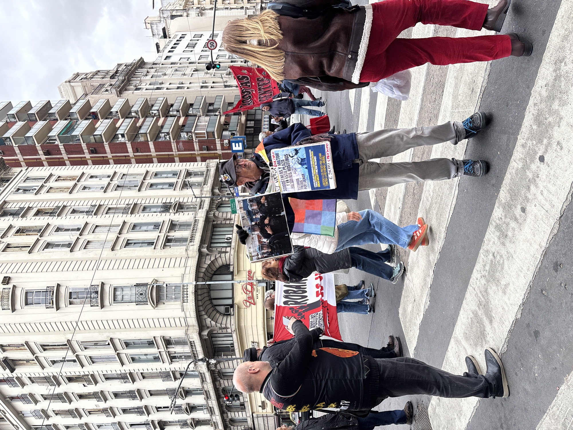 The image shows a group of people protesting in the city of Buenos Aires, Argentina. They are standing in a crosswalk on a street lined with tall buildings, one of which has a La Opera sign. The protesters are holding signs and banners with messages written in Spanish. One sign reads "SOMOS ENEAS Y MILTON" and "ESTAMOS PRESOS POR LUCHAR" (We are Eneas and Milton, We are prisoners for fighting). Another banner says "ROBAR". Some of the protesters are wearing casual clothing, while others are dressed in more formal attire. The weather appears to be overcast. A traffic sign indicating a speed limit of 30 is also visible.