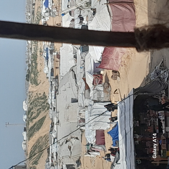 An aerial-level view captures a densely packed displacement camp in Khan Yunis, Palestinian Territory, under a hazy sky. The foreground features a rudimentary market stall on the left, with goods displayed under a white tarpaulin roof. A person in dark attire is partially visible near the stall. A thick, dark wooden post or beam obscures the right side of the frame. A thin, dark cable traverses diagonally from the upper left to the lower center.
The midground is dominated by an expanse of makeshift shelters and tents constructed from various materials, including white, grey, blue, and maroon tarpaulins, set on sandy ground. Several individuals are present: a small group gathers near a blue tent on the left, while two individuals in dark clothing walk towards the left in the center, and another moves right. Some shelters appear partially damaged or collapsed.
In the background, a large sandy embankment with sparse green vegetation rises, topped with additional scattered tents. A slender communication tower is visible on the horizon to the left. The sky is light and mostly clear, with a noticeable haze.