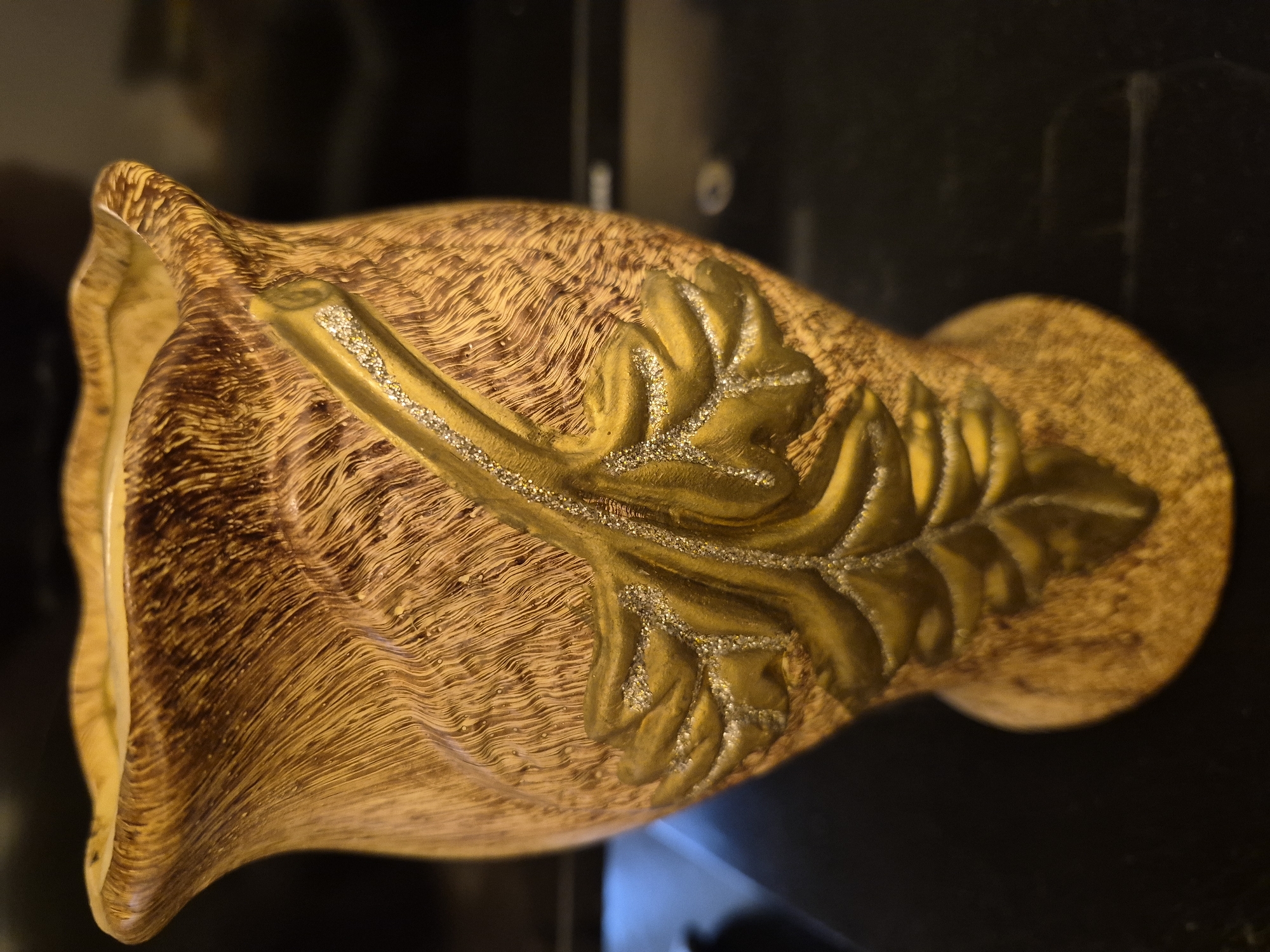 A close-up, eye-level shot showcases a decorative ceramic vase with a textured, wood-grain-like finish in shades of brown and gold. A raised, golden leaf design with glitter accents adorns the front of the vase. The vase has a fluted rim and a rounded base. The background is dark and out of focus, suggesting an indoor setting, possibly a shelf or table. No specific location in Vanadzor, Armenia, can be determined from the image.