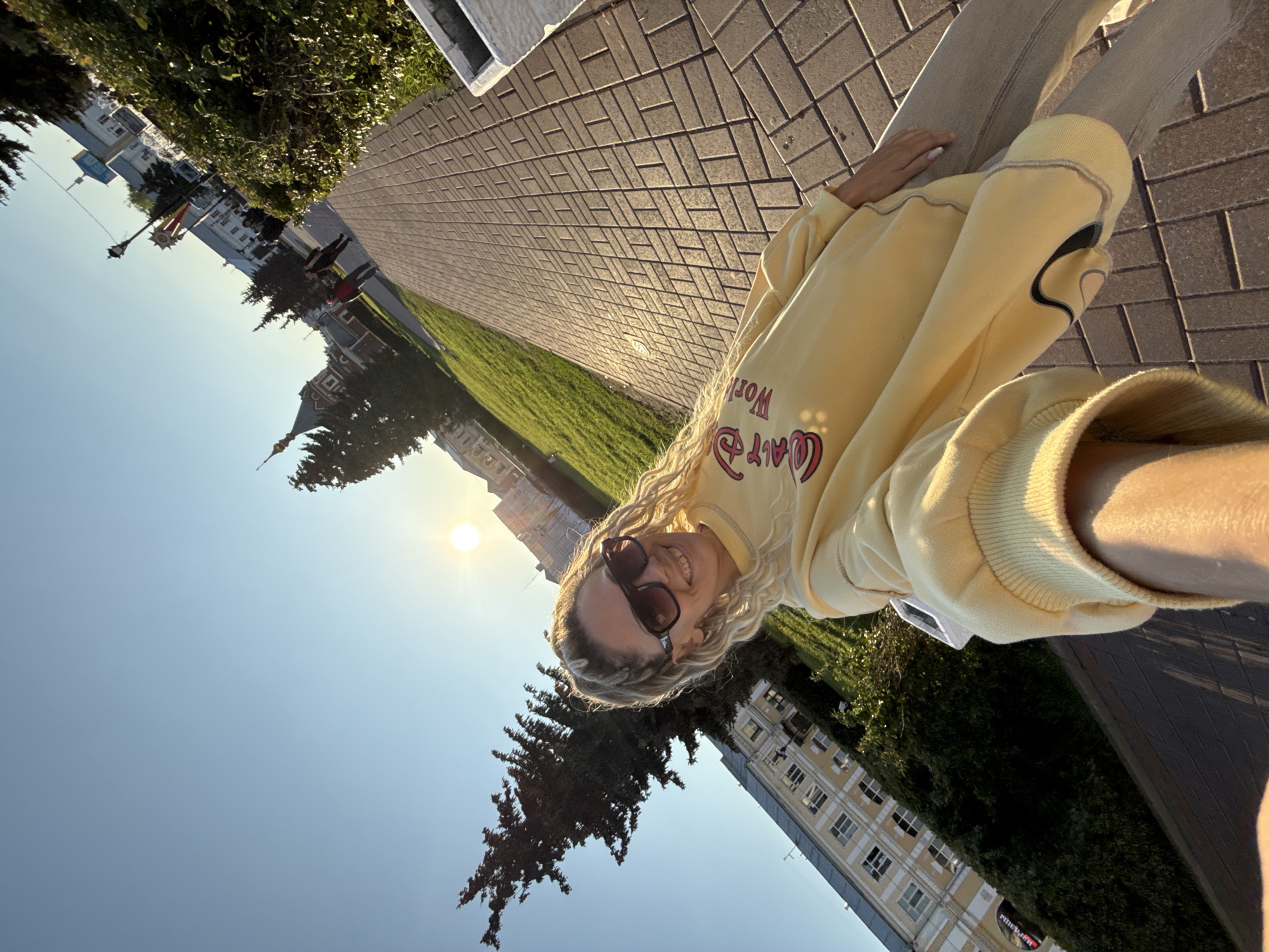A woman with blond curly hair and sunglasses smiles into the camera. She is wearing a yellow sweatshirt and light-colored pants. The picture appears to be a selfie taken in Шебунино, Russia. Buildings and trees can be seen in the background, as well as a brick walkway and a grassy area. The sun is shining brightly in the sky.