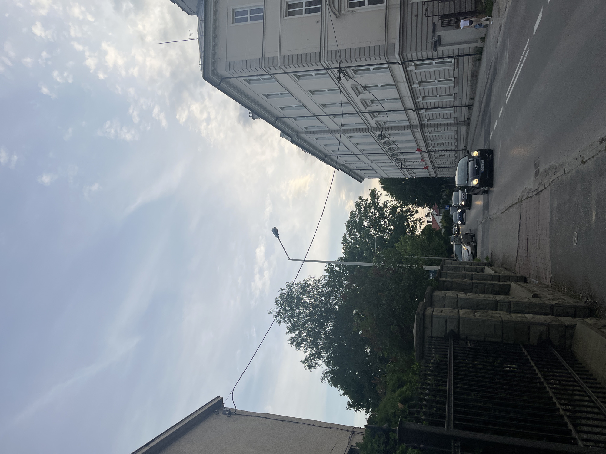 The image captures a street scene in what appears to be an urban or suburban environment, viewed from a slightly elevated perspective.
**Subjects:**
* **People:** One individual wearing a white shirt is partially visible near the entrance of the larger building on the right side of the street.
* **Objects:** Several cars are present on the road, including a dark-colored sedan in the foreground with its headlights illuminated, and other vehicles queuing behind it. A tall street lamp extends over the trees in the mid-ground. On the right, a stone wall topped with a black metal fence runs alongside the street. Utility wires or cables crisscross the scene, connecting to buildings and poles. A small antenna is visible on the roof of the prominent building.
* **Scene:** The setting features a two-lane asphalt road with double white lines, bordered by a sidewalk on the right. A large, multi-story building with numerous windows and architectural detailing dominates the right side of the frame. A simpler, smaller building is partially visible on the left. Dense green trees fill the mid-ground and parts of the foreground.
* **Setting:** A street lined with buildings and mature trees.
**Activity or event taking place:**
Cars are either driving slowly or stopped in traffic on the road, indicative of typical daily commute or movement.
**Notable details:**
The time of day appears to be late afternoon or early evening, suggested by the soft light and the fact that some car headlights are on. The sky is partly cloudy, with scattered white clouds against a lighter background. Architectural styles, particularly of the larger building, and visible vehicle license plate prefixes (e.g., "S2A" and "DW") suggest a European, likely Polish, location.
**Any visible text:**
A partial license plate "S2A 77CS" is visible on the front of the dark sedan. Another license plate further down the road begins with "DW".