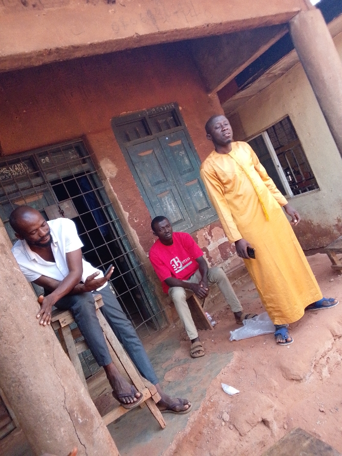 Three adult males are observed in an outdoor setting in Ankpa, Nigeria, against a reddish-brown building with an overhanging eave. The ground is reddish-brown earth.
The individual on the left, a dark-complexioned male with a beard, is seated on a wooden stool. He wears a white collared shirt, dark trousers, and dark flip-flops. He holds a black smartphone in his left hand, looking downwards at it.
The individual in the center, a dark-complexioned male, is seated on a wooden stool. He wears a red short-sleeved t-shirt with a white logo and text, and khaki-colored trousers. He looks towards the right. He wears brown sandals.
The individual on the right, a dark-complexioned male with short hair, stands upright. He wears a long, flowing yellow garment and blue flip-flops. He holds a black smartphone in his right hand and looks upwards with his eyes closed or partially closed.
The background features a building wall of reddish-brown material. To the left, a window with a metal security grille is visible. Markings, including "MB (ABI)" and numbers "2034 240," are on the wall adjacent to this window. A green or dark blue wooden double door is situated centrally in the background. To the right of the door, another window opening is present. On the ground, near the feet of the rightmost individual, a transparent plastic bag containing a white object and a separate small white piece of debris are visible.