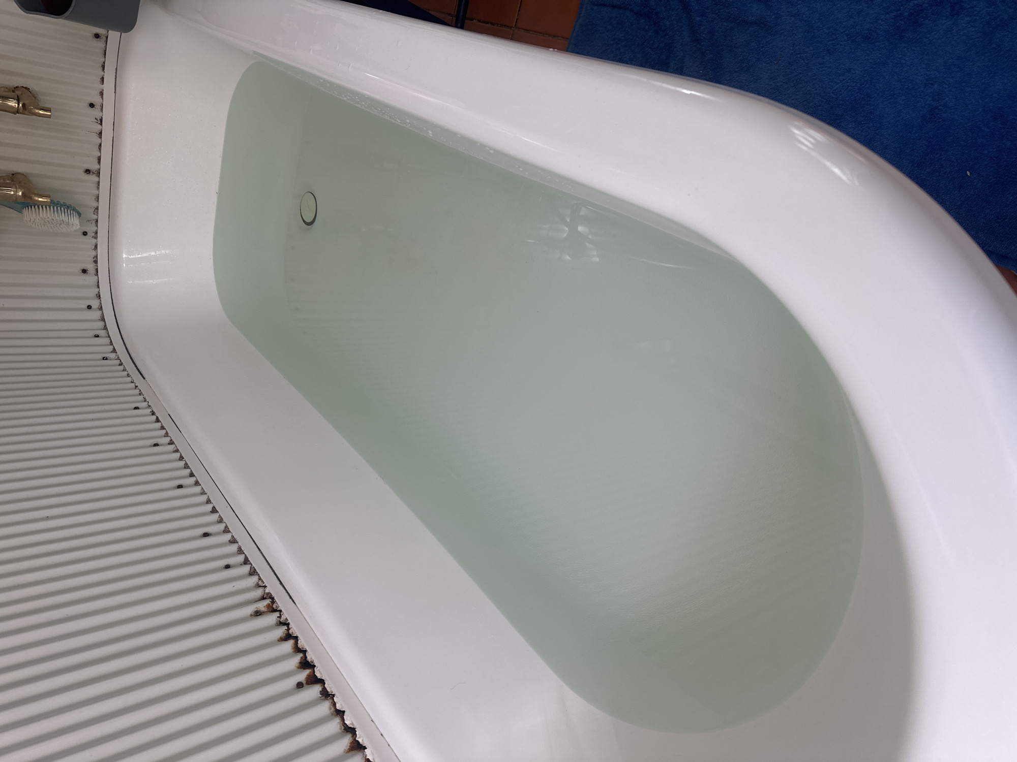 A bathtub filled with clear water is depicted. The tub is white and appears to be made of porcelain. The walls surrounding the bathtub are made of corrugated metal and show signs of rust. There are brass fixtures and a blue brush visible above the tub. A dark blue towel or mat is visible on the floor to the right of the bathtub. The image appears to be taken indoors, likely in a bathroom. The location is Thora, Australia.