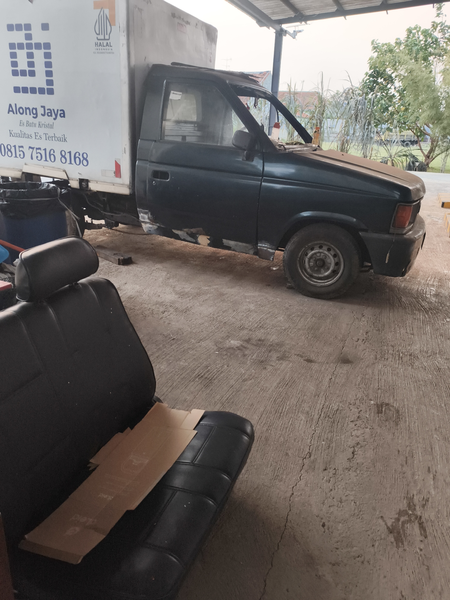 The image shows a dark green pickup truck with a white cargo box in what appears to be a repair shop or garage in Pekalongan, Indonesia. The truck's bodywork, particularly the lower panels, shows signs of repair or modification, with some sections appearing unfinished or replaced. The cargo box is branded with "Along Jaya" and text that translates to "Crystal Ice" and "Best Ice Quality" along with a phone number. A "HALAL INDONESIA" certification is also visible on the cargo box. In the foreground, a black car seat is placed on the concrete floor, with some cardboard pieces on it. The environment suggests an outdoor or semi-covered workspace, with a concrete floor and some greenery and structures visible in the background. The lighting indicates it might be late afternoon or early morning. There are no people visible in the image.