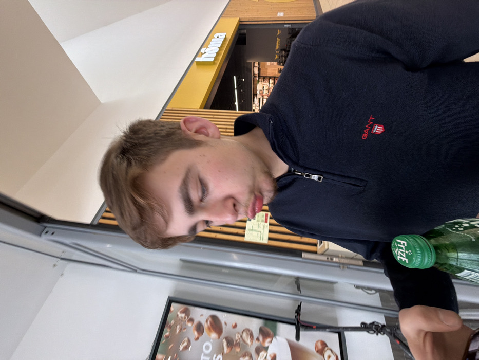 A young male, estimated to be in his late teens or early twenties, is depicted from the mid-chest upwards in an indoor commercial setting, likely a shopping mall in Vila Real, Portugal. He has short, light brown hair and a light complexion, presenting a pouting facial expression with puckered lips while looking downwards and to his right. He is wearing a navy blue quarter-zip sweater with a small red and white GANT shield logo on the left chest. His left hand is visible in the lower left foreground, holding a green plastic bottle with a matching green cap, clearly marked with the inverted white text "Frize."
In the background to the left, a portion of a large vertical display or poster is visible, advertising a beverage with scattered hazelnuts and partial text. Below this display, the handlebars and front wheel of a black and silver electric scooter are present. Directly behind the male subject, a wall features vertical light-toned wooden slats. To the right of the subject, a retail storefront is visible, identified by a prominent yellow sign displaying "hômə" in white lettering, with merchandise visible within the store's interior. The ceiling above is white and angled, indicative of modern commercial architecture, and the area is brightly lit by overhead lighting.
