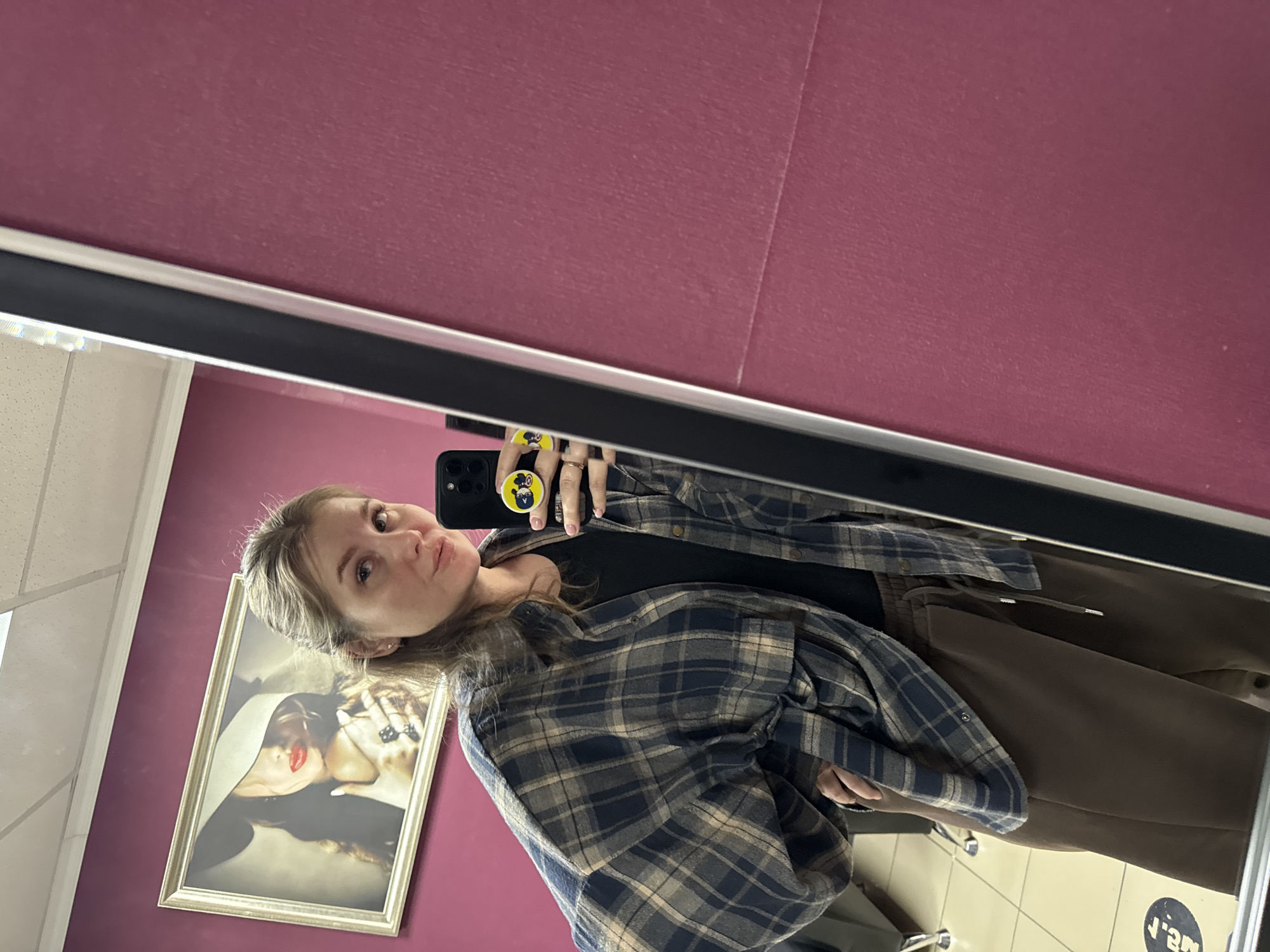 A young woman is taking a selfie in a mirror. She is wearing a plaid shirt over a black top, and brown pants. The mirror is in a room with magenta walls. On one wall is a framed portrait of a woman in a hat. The floor has tiles with a blue circle and text on them. The photograph appears to be taken in Perm, Russia.