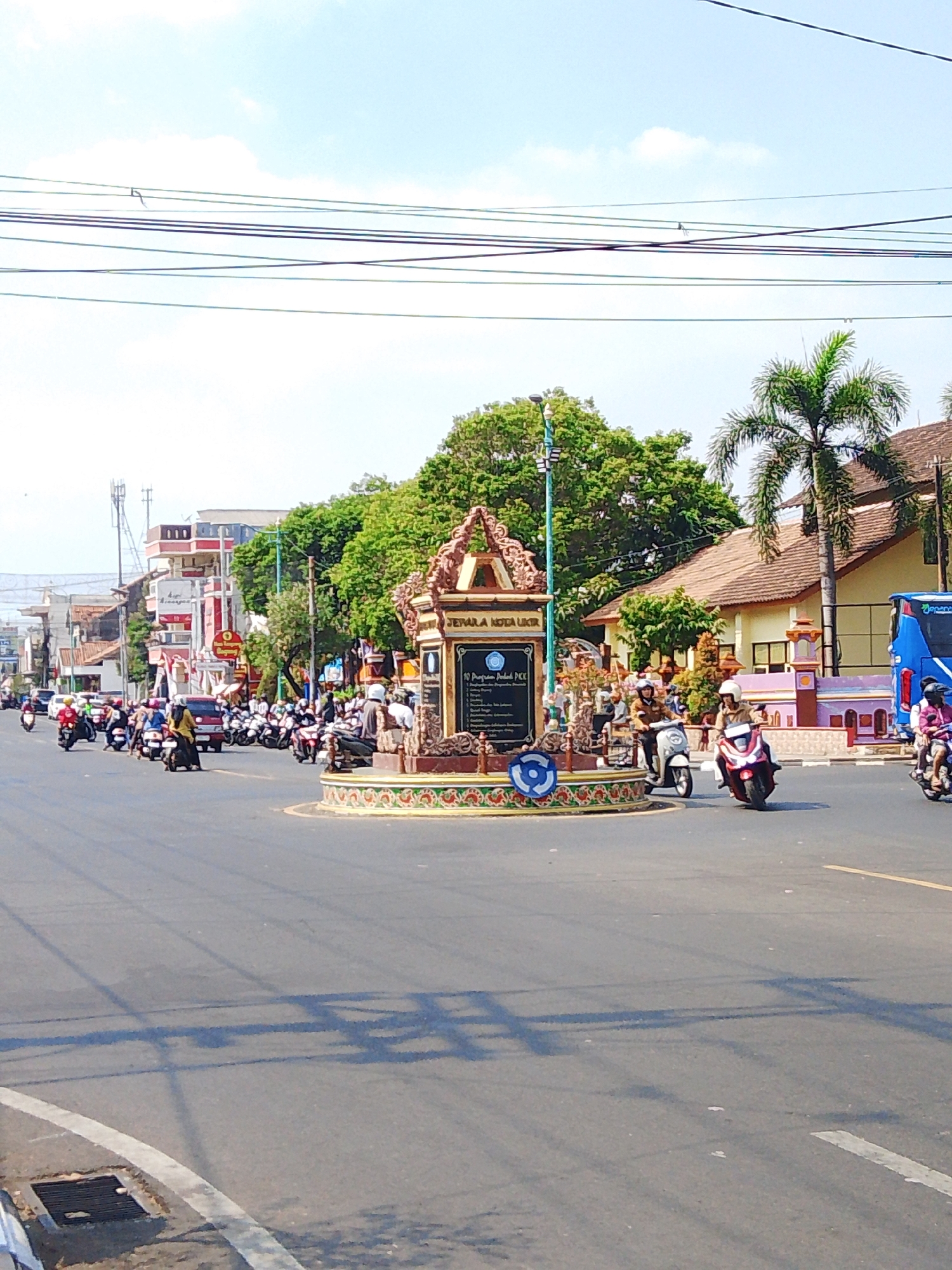 This is a medium-shot, eye-level, outdoor photograph of a busy intersection in Jepara, Indonesia. The image is dominated by a traffic circle in the foreground with a decorative monument at its center. The monument is elaborately decorated with what appears to be a traditional Javanese architectural style, featuring carved details and a colorful base. Text is visible on the monument, suggesting it is informative or commemorative.
The traffic circle is surrounded by a paved road with white lane markings. Numerous motorcycles and a few cars are navigating the intersection, indicating a moderate level of traffic activity. Many individuals are riding on motorcycles, some wearing helmets. A large blue bus is visible on the right side of the frame, alongside a building with a brown tiled roof. Behind the traffic circle, a street lined with buildings and trees extends into the background. The buildings appear to be a mix of commercial establishments, with some featuring red and white signage. A tall antenna or cell tower is also visible in the distance. The sky is clear with a few scattered clouds, and the overall lighting suggests it is daytime. The presence of overhead power lines indicates an urban or semi-urban setting. The photograph captures a slice of daily life at a public intersection in Jepara.