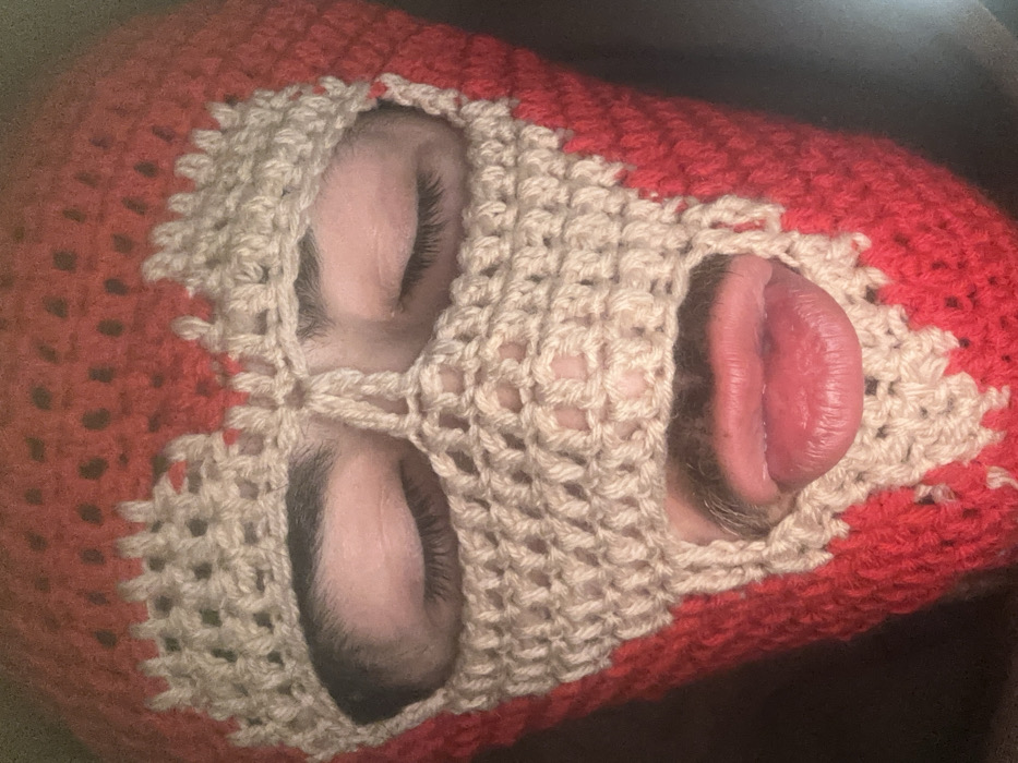 A person is depicted in a close-up, head-on view, wearing a two-toned, crocheted balaclava. The balaclava is predominantly red, with a beige or light tan pattern on the front around the facial apertures. This beige pattern forms jagged, irregular outlines around the eye openings, suggestive of a broken crown or heart shape above the eyes, and a lattice-like covering over the nose and upper mouth area. The person's eyes are closed, showing dark eyebrows and eyelashes. Below the beige crochet work covering the nose and upper lip, the person's mouth is open, with the tongue extended. Full, reddish lips are visible, along with faint facial hair around the mouth opening. The background is dark and out of focus, offering no discernible environmental details. The location in the city of Carnin, France, cannot be determined from the visual information present in the image.