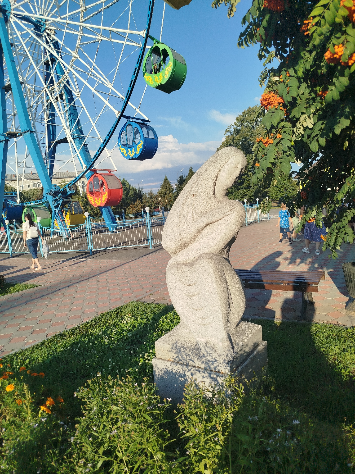 The image captures a sunny day in an amusement park in Yalutorovsk, Russia. In the foreground, a stone sculpture of a woman in a kneeling pose is illuminated by the sun, casting a shadow on the paved walkway behind her. Green grass and flowering plants surround the sculpture. To the left, a large blue Ferris wheel dominates the skyline, with colorful cabins – green, blue, and red – suspended from its structure. A woman with a white backpack walks along the patterned brick path, moving away from the camera. In the background, trees and buildings are visible under a clear blue sky with scattered clouds. Several children are also seen in the middle ground, walking on the path near a wooden bench. The overall atmosphere suggests a public recreational space on a pleasant day.