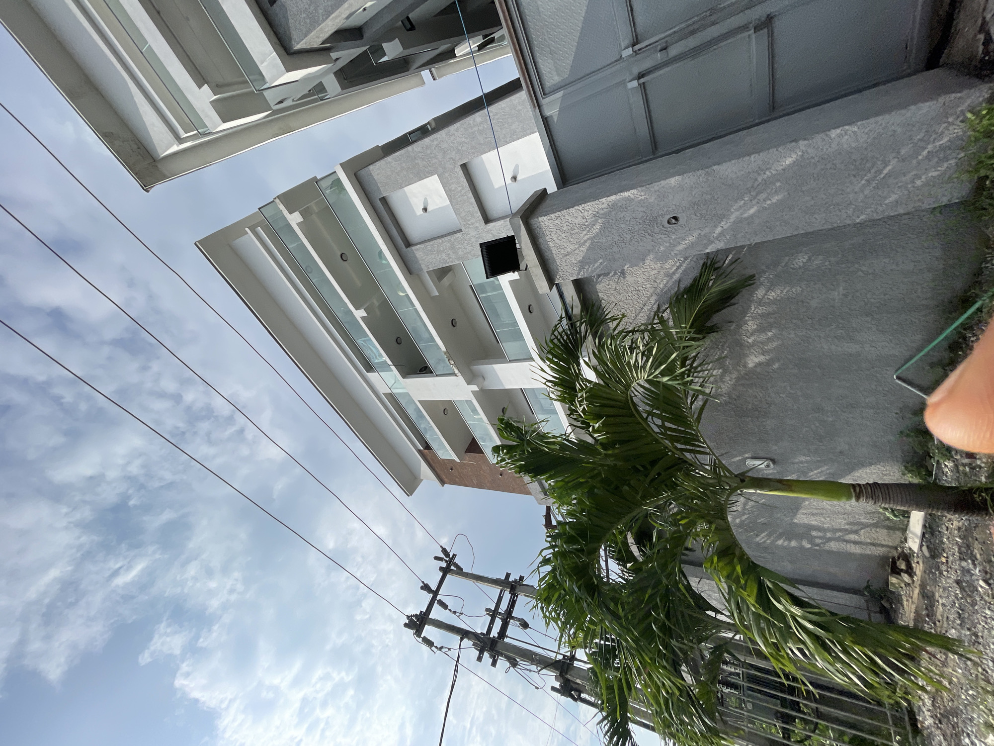 The image shows a multi-story building with balconies and glass railings, located in Ilado, Nigeria. A palm tree is in the foreground, and power lines are visible against a cloudy sky. The building appears modern, with a combination of white and grey facade. The overall impression is an urban landscape with a touch of tropical vegetation.
