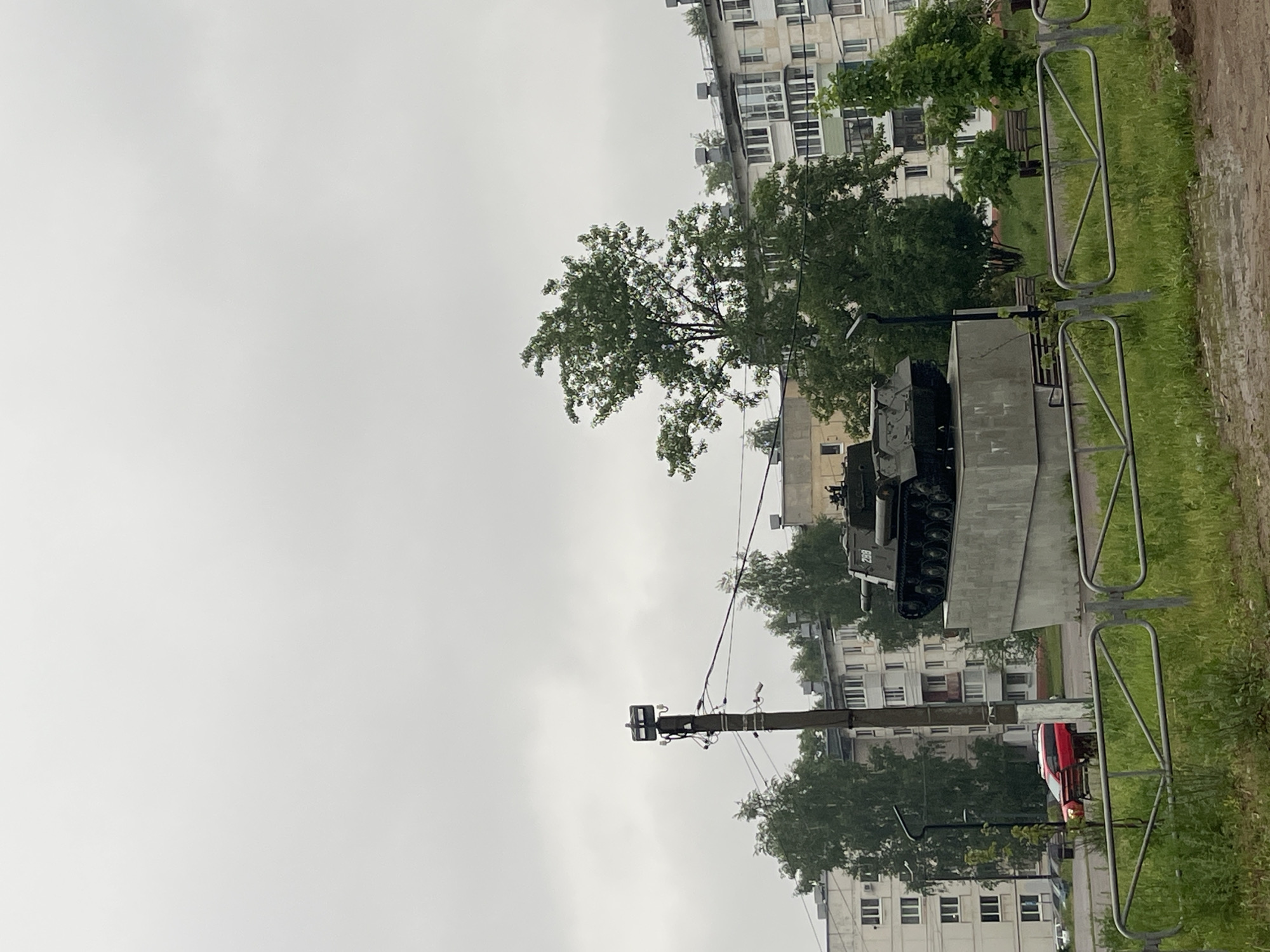 The image depicts a tank monument in the city of Chaikovsky, Russia. The tank is mounted on a concrete plinth and is surrounded by metal fencing. Behind the monument are apartment buildings and trees. The sky is overcast. A utility pole is visible in the foreground. A red car is parked near the monument.