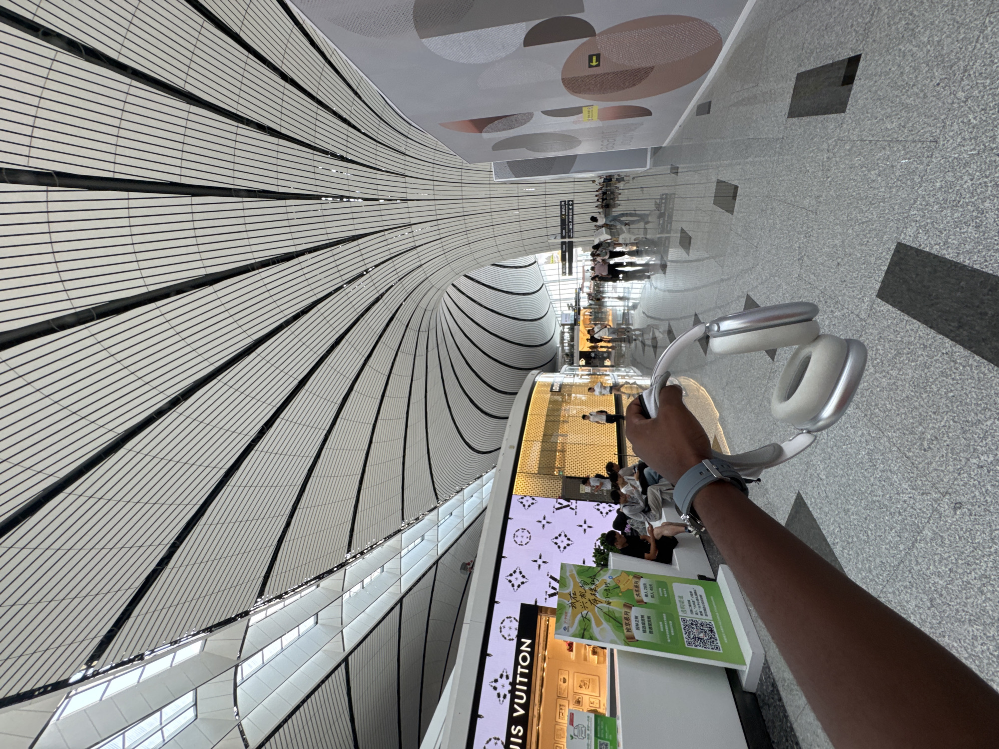 A person's arm extends into the frame from the left, holding a pair of white headphones. The arm is dark-skinned and wears a grey watch. The background depicts the interior of a modern airport terminal. The ceiling is a prominent architectural feature, with white panels and black beams forming a sweeping, curved structure. Large windows and skylights allow natural light to illuminate the space.
To the left, a section of a Louis Vuitton store is visible, with displays of merchandise and a large screen showing the brand's logo and patterns. Beside the store, several people are seated, appearing to be waiting.
Further into the terminal, a polished, reflective floor stretches into the distance. Other travelers are walking and standing in various parts of the concourse. A large, abstract mural with circular shapes in shades of grey, brown, and pink is mounted on a wall to the right. Directional signs are also visible in the distance. The overall impression is one of a spacious, contemporary, and well-lit transportation hub, likely Beijing Daxing International Airport in Guangyang District, China, given the architectural style and presence of luxury retail.