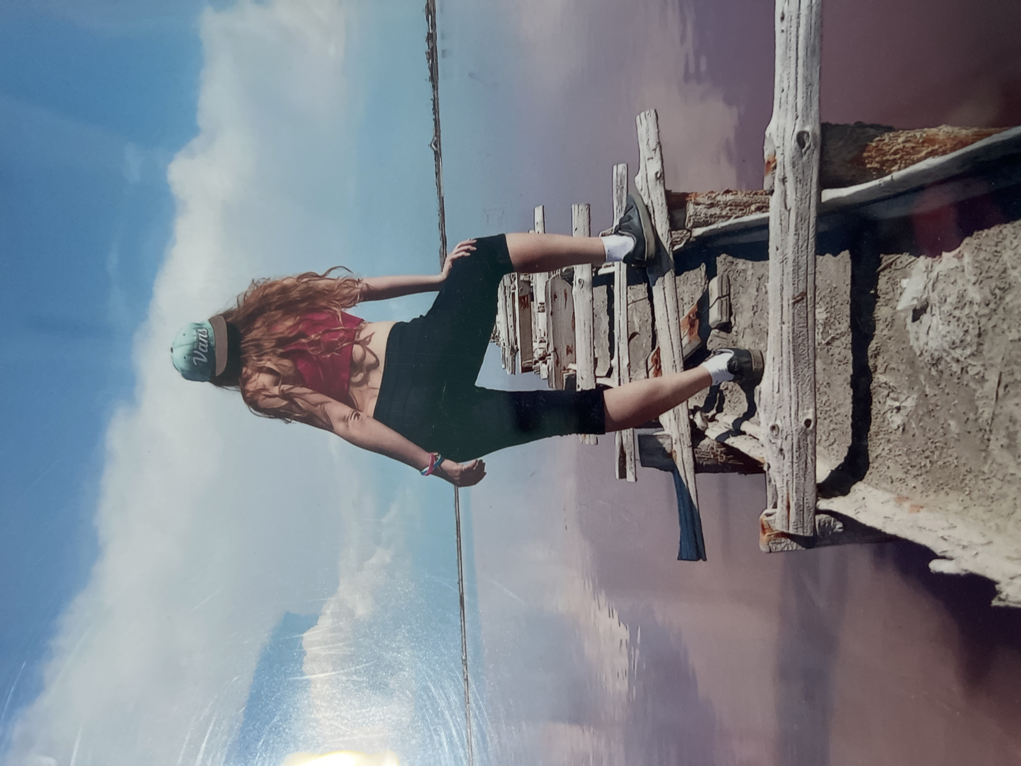 A young woman stands on a wooden structure with a pink, reflective body of water and a blue sky with clouds in the background. She wears a red top, black shorts, a blue cap with "Vans" on it, and white socks with shoes. The wooden structure appears aged and weathered. The image is possibly taken at Lake Sasyk-Sivash, near Yevpatoriya, Ukraine, known for its pink waters and wooden structures.