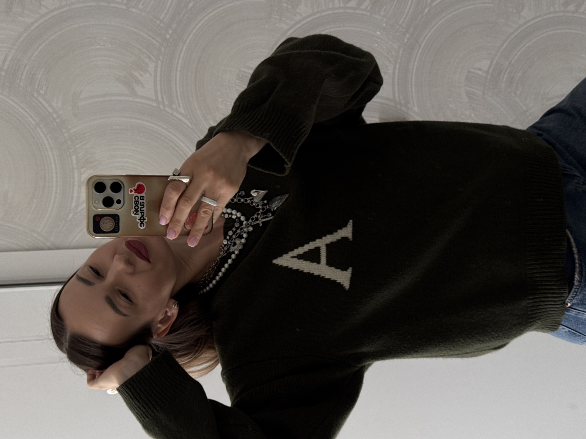 A woman is taking a selfie indoors. She has her head resting on her hand and is holding her phone up to take the picture. She is wearing a dark green sweater with a white "A" on the front, jeans, and a necklace. The phone has a sticker on the back. The background is a wall with a patterned design. The location cannot be determined as Sochi, Russia.