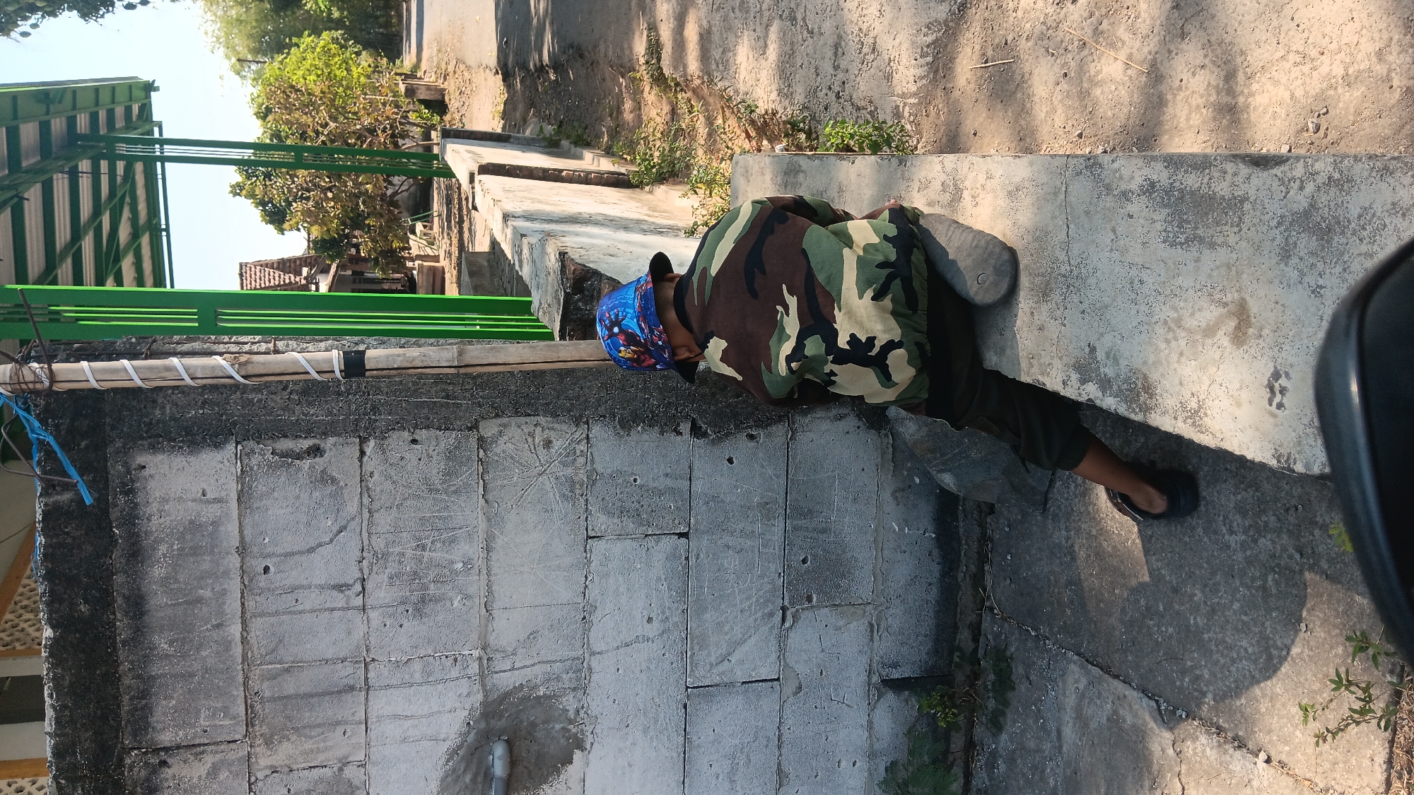 In Gemenggeng, Indonesia, a young child wearing a camouflage jacket and a blue hat with a superhero motif is seated on a concrete ledge. The child is facing away from the camera, towards a rough, textured grey brick wall. The environment appears to be outdoors, with a dirt path and vegetation visible in the background. A green metal structure with a corrugated roof is also present, suggesting a shelter or building addition. The lighting indicates it is daytime, with shadows cast by the structures and the child. The overall scene is casual and possibly depicts a child resting or observing their surroundings.