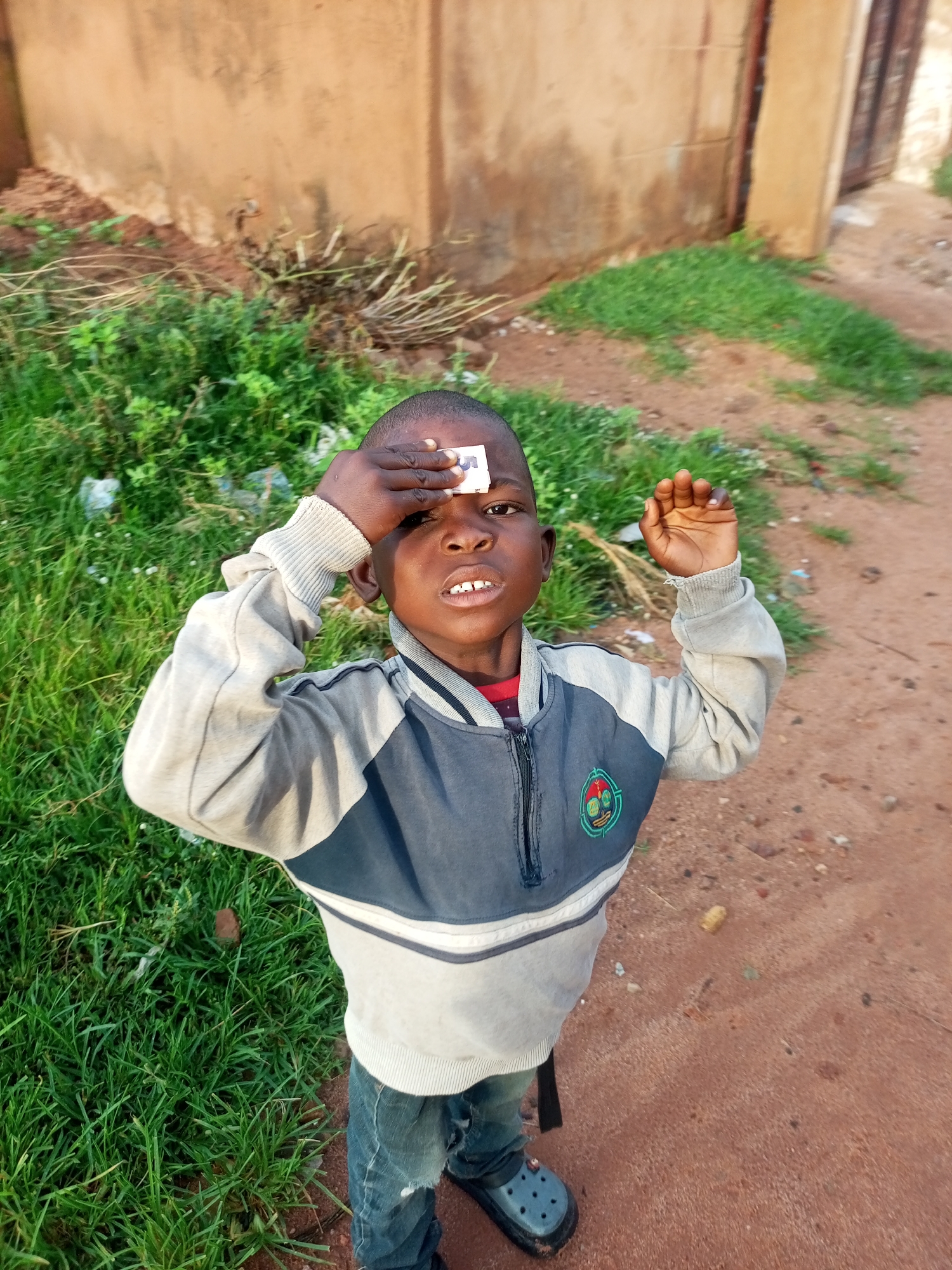 A young boy is standing outdoors, with a blurred background of a building and vegetation. He is wearing a grey and dark blue zip-up jacket over a red shirt, and blue jeans. A white card or sticker is placed on his forehead. His right hand is raised to his forehead, partially obscuring the object. He appears to be looking towards the camera with his mouth open. The ground is a mix of reddish-brown dirt and green grass. The scene is likely in a rural or semi-urban area within Tempure, Nigeria, given the visible environmental cues.