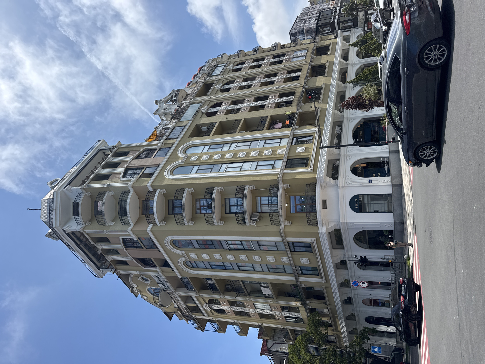 The image shows a tall, beige building in Batumi, Georgia. The building has multiple stories, featuring arched windows, balconies with black metal railings, and ornate white trim. The sky is blue with scattered white clouds and a contrail. In the foreground, cars are parked on the street. One individual is visible on the sidewalk near the building's entrance. A traffic sign is also visible.