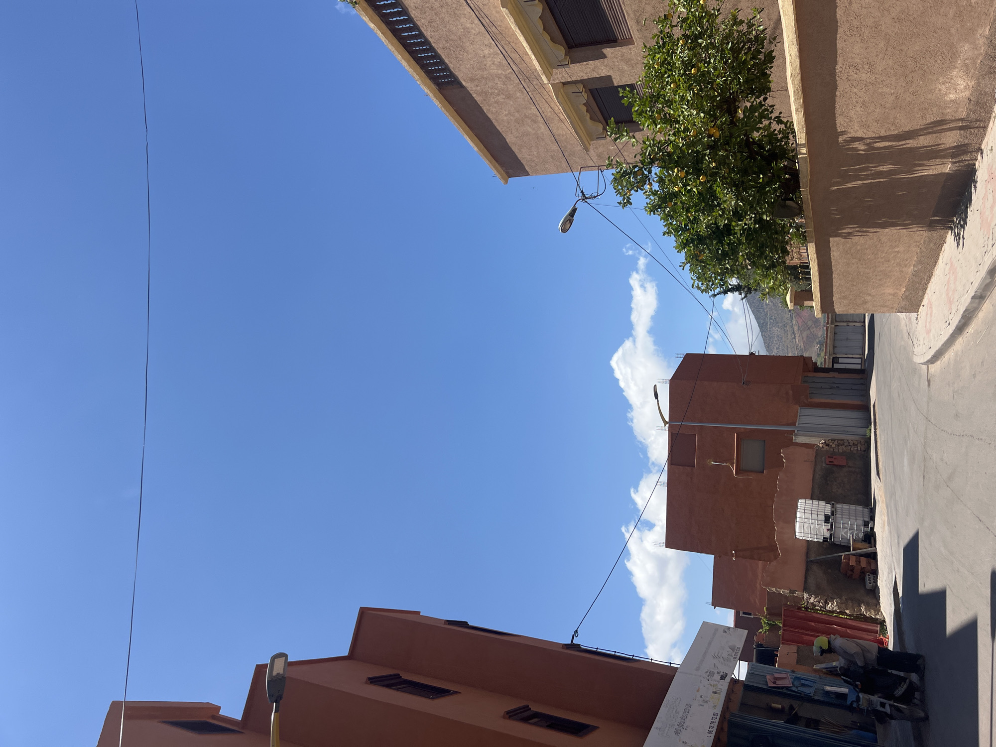 The image, taken in Ouaouizert, Morocco, shows a street scene under a bright blue sky with scattered clouds. Buildings line the street, constructed in earth-toned hues. Power lines stretch across the sky between the structures. A tree with green foliage and what appear to be citrus fruits grows near the top right building. A partially visible mountain can be seen in the distance.
On the right side of the street, a person in a jacket and helmet stands near a parked motorcycle. A stack of rectangular containers and construction materials are present nearby. A sign with lettering is partly visible near the bottom of the image. The street pavement shows visible wear and tear.