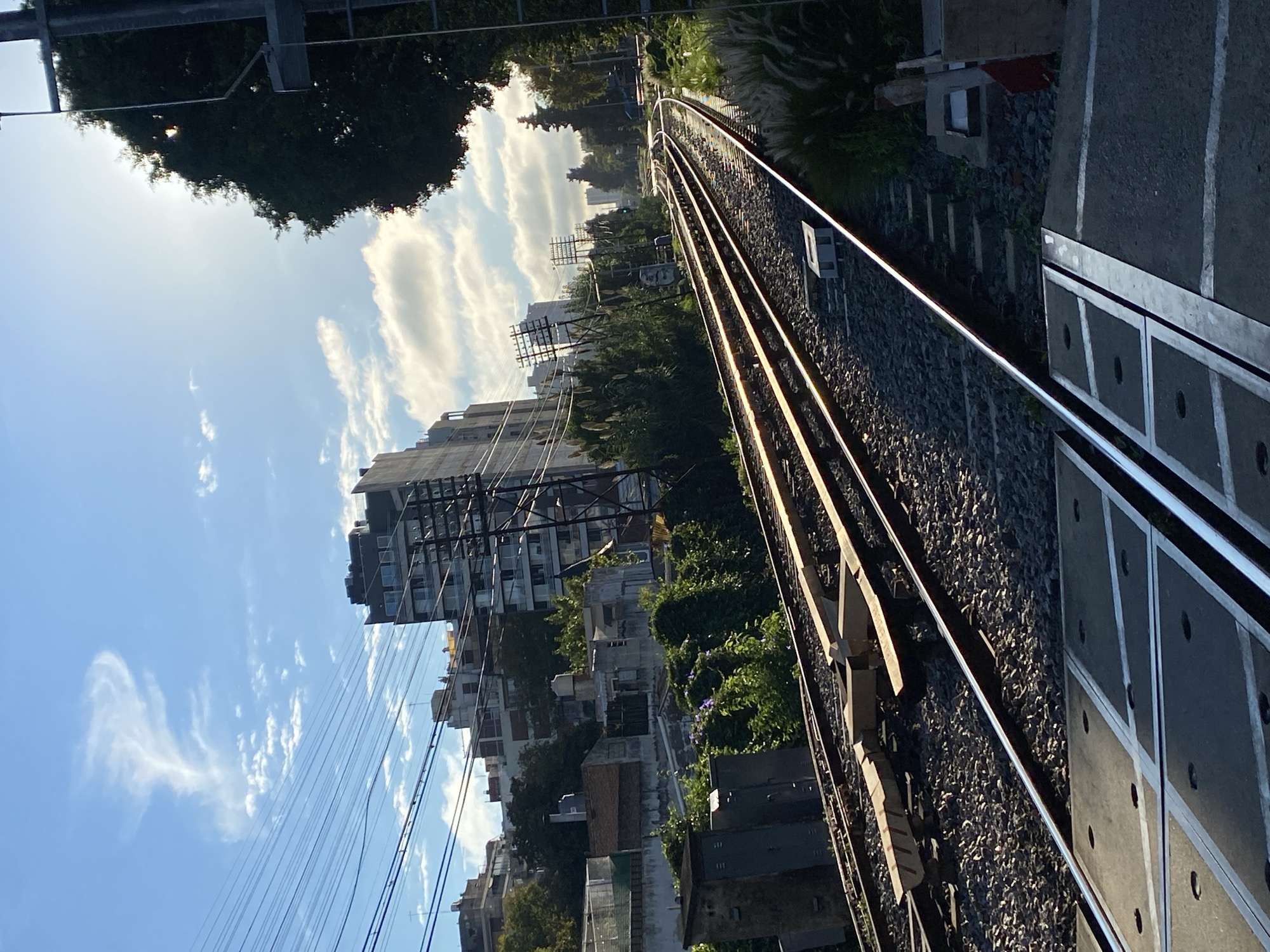 The image depicts a railway track in Buenos Aires, Argentina. The tracks run diagonally through the frame, with the perspective suggesting a view from above. The right side of the frame shows a concrete platform with embedded metal strips and circular holes. To the left of the tracks, there is crushed rock ballast. Beyond the tracks, urban buildings of varying heights are visible, including apartment complexes, smaller houses, and vegetation. Overhead power lines and utility poles cut across the sky. The sky is light blue with scattered cumulus clouds. Bright sunlight illuminates the scene.