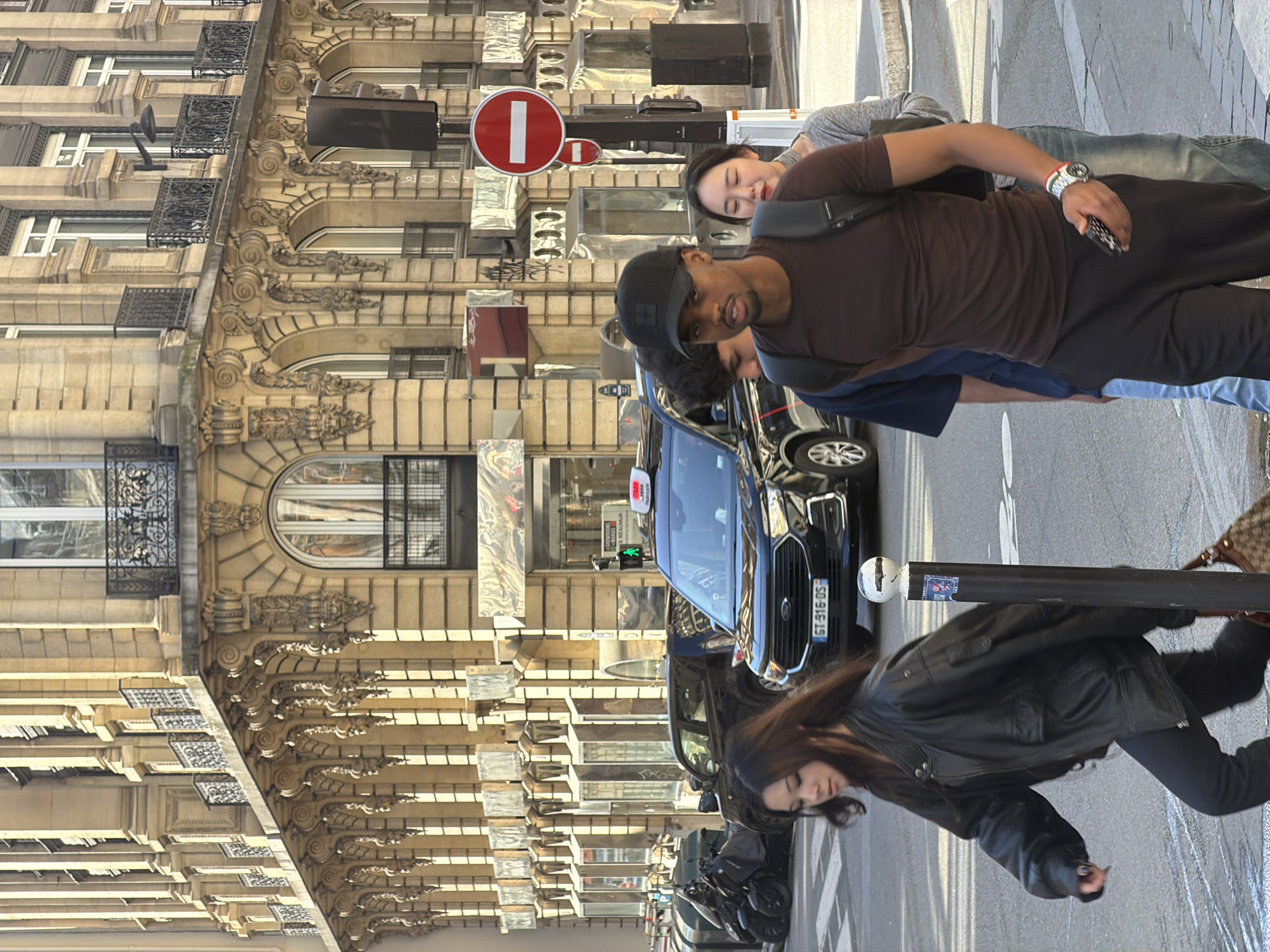 The image shows a street scene in Paris, France. The architecture of the buildings suggests a classic Parisian style, with decorative moldings, balconies, and tall windows.
Several people are present:
* A man wearing a black baseball cap and a brown shirt is in the foreground. He is carrying a backpack and appears to be standing.
* A woman is partially visible behind the man.
* Another woman is walking on the sidewalk, wearing a black leather jacket and black pants.
A black taxi is stopped on the street, and motorbikes are parked.
A no entry sign is visible.
The environment suggests a typical Parisian street with a mix of pedestrians and vehicles. The weather appears sunny.