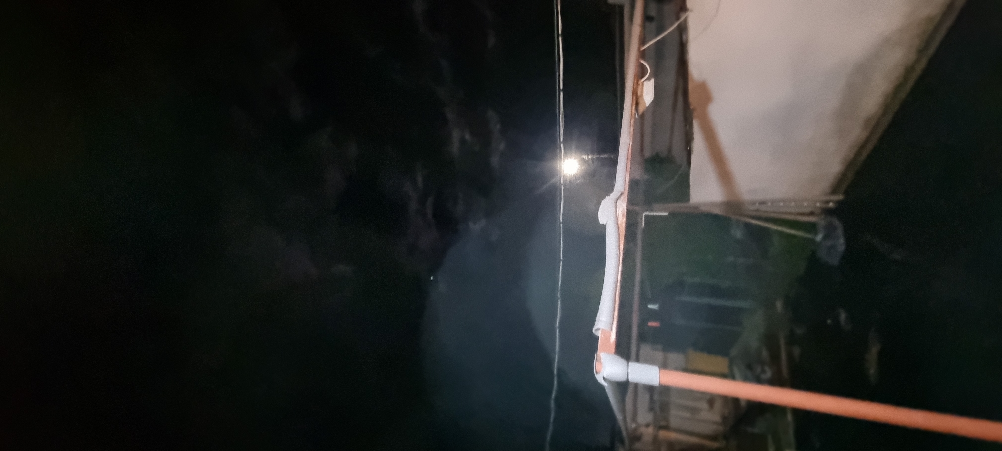 The image is taken at night in an outdoor environment. The scene features architectural structures, including a building with a light-colored wall and a structure resembling a roof or awning made of a lighter material. An orange PVC pipe or conduit is visible in the foreground, possibly part of an electrical or plumbing system. Other details include wiring, supports, and what appears to be vegetation or terrain in the background, which has very dark shades. A light source is seen in the distance, possibly a streetlight. There is no indication of specific objects or people visible. The location is in the city of Lom, Bulgaria.