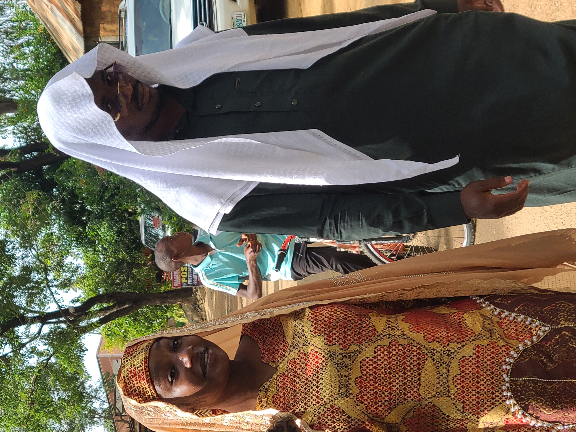 Two individuals, a woman and a man, stand outdoors in what appears to be a street scene in Jos, Nigeria. The woman, positioned on the left, is wearing a traditional African dress and headscarf. The dress is richly patterned with gold and red, and adorned with white pearls along the hem. Her headscarf is a light beige color, and she is also wearing earrings. She is looking towards the camera with a slight smile.
The man, on the right, is wearing a dark green robe with a white head covering. He has glasses on and is looking downwards and to his left. A white vehicle with a license plate visible, displaying "94CU," is parked behind him. In the middle ground, a young boy in a light blue t-shirt and dark pants is visible. He is standing near a bicycle. The background shows trees and a building with a corrugated iron roof, consistent with the architecture found in Jos. The overall impression is a snapshot of daily life in a Nigerian city.