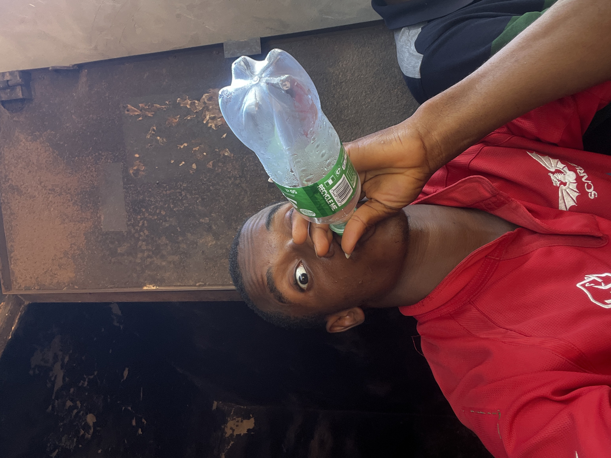An image, oriented vertically, shows a young Black man drinking from a clear plastic water bottle. He is positioned next to what appears to be a large, dark, weathered metal or concrete container, possibly a refuse bin or a deep industrial structure, with visible rust and peeling textures on its inner walls. The man is wearing a bright red short-sleeved shirt featuring a white logo of a winged creature (resembling a dragon or gargoyle) and partially visible text that reads "SCARF" next to it. He also has dark trousers with a grey and green stripe visible.
His left arm is raised, holding the bottle to his lips, and condensation is visible on the bottle's exterior. His head is tilted slightly upwards, and his right eye is wide open, looking directly towards the camera, conveying a somewhat surprised or direct gaze. The background behind him and to his left is dark and indistinct, suggesting an enclosed or deeply shaded area.
Visible text on the water bottle's green label includes "RECYCLE ME" and a barcode, along with "50cl" and other smaller, unreadable text. The scene likely takes place in an urban or industrial part of Onitsha, Nigeria, given the provided context and the utilitarian appearance of the surroundings. The time of day is not explicitly clear, but there is enough light to illuminate the subject and the bottle.
