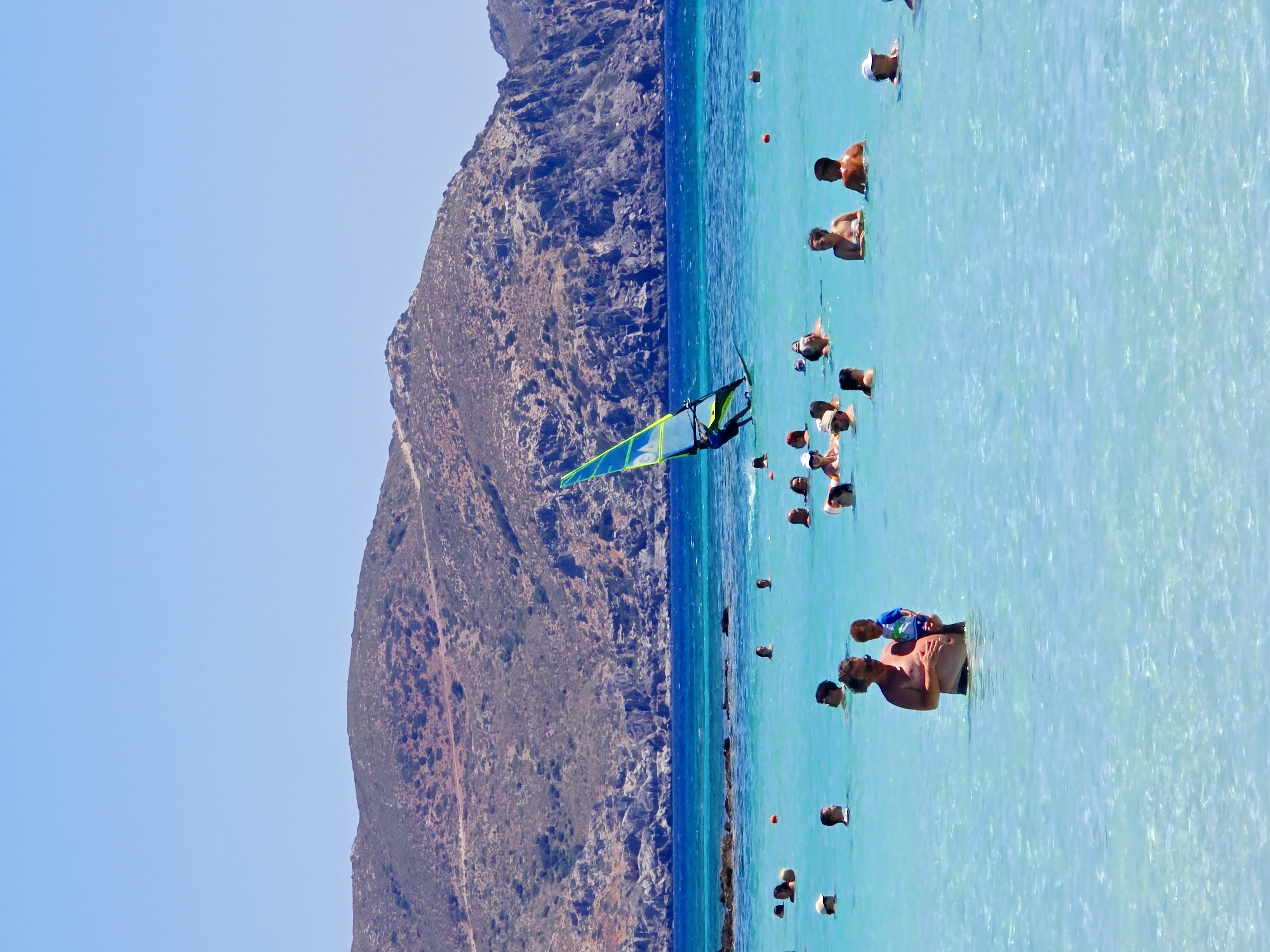 The image depicts a crowded beach scene in Greece, under a clear blue sky. In the foreground, a man is standing in shallow, clear turquoise water, holding a young child. The water is populated by numerous other swimmers, their heads and shoulders visible above the surface. Further out in the water, a person is windsurfing, their sail a vibrant green and yellow against the blue expanse. In the background, rugged, brown hills with sparse vegetation rise from the sea. The overall impression is a lively and sunny day at a popular coastal destination.