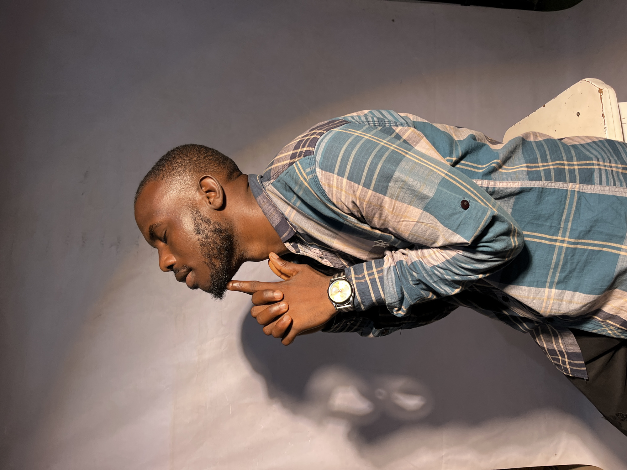 A medium shot shows a Black man from the chest up, seated and facing to the left, with his eyes closed and head tilted back. He has short hair and a goatee. His hands are clasped in front of his face, with his right index finger touching his chin. He is wearing a plaid shirt with blue, tan, and white checkered patterns. A wristwatch with a silver band and white face is on his left wrist. He is sitting on a white stool with visible wear and tear. The background is a plain gray backdrop with a soft shadow of the man on the left side. The lighting appears to be coming from the left, illuminating the man's face and right side.