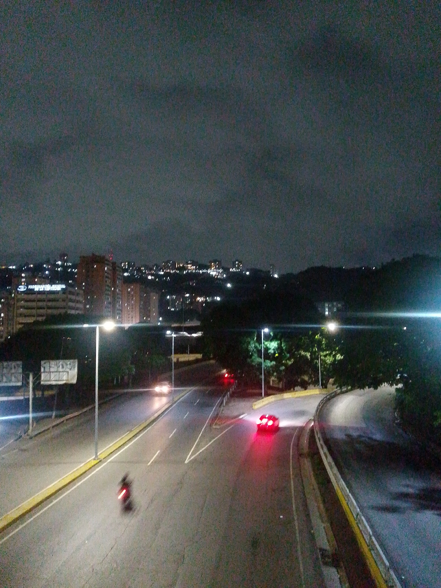 The image depicts a nighttime urban scene in Caracas, Venezuela. The photograph is taken from an elevated position, likely an overpass or building, overlooking a multi-lane highway. The city skyline is visible in the background with scattered lights from buildings and residences. The sky is overcast and dark.
On the highway, a motorcycle is visible traveling in the left lane. Further ahead, a red car is moving on the right side of the road. Streetlights illuminate the highway, casting a bright glow. The road surface shows signs of wear and tear. There are guardrails visible on the right side of the highway.
The presence of illuminated buildings suggests the location is likely within the metropolitan area of Caracas, with a mix of residential and commercial properties in the vicinity. The hilly terrain is visible on the right, indicating that the location might be close to the mountainous areas surrounding the city.