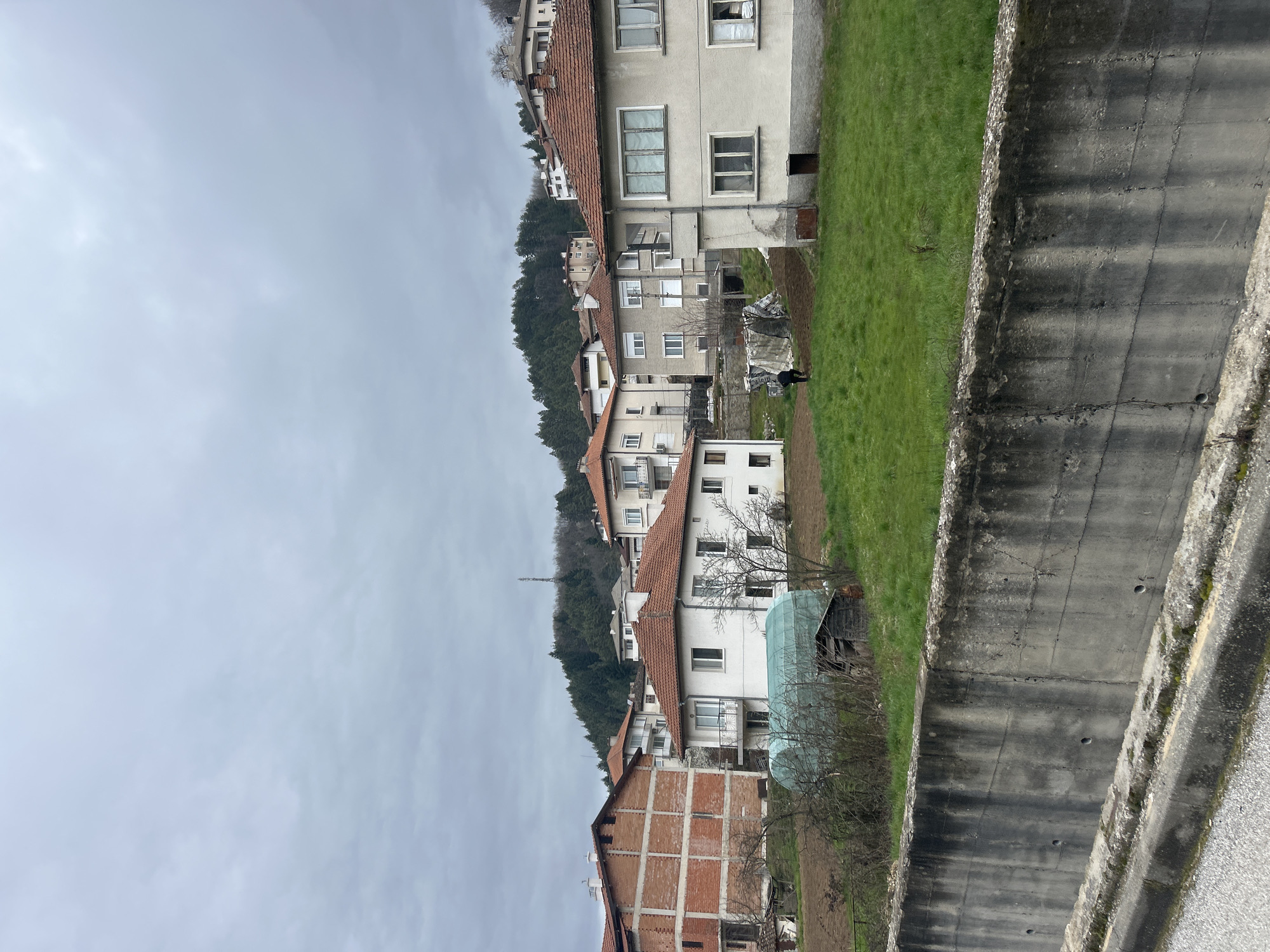 The image shows a residential area in Zlatograd, Bulgaria, with houses lining a hillside. A concrete wall runs along the right side of the image, likely a retaining wall for a road or path. The houses vary in style, with some having red tile roofs and others appearing to be made of brick. There is green grass and patches of bare earth between the houses. The sky is overcast. A communications tower is visible in the background on the hillside. A person is standing in the grass between two houses. There appears to be a small greenhouse in one of the yards.