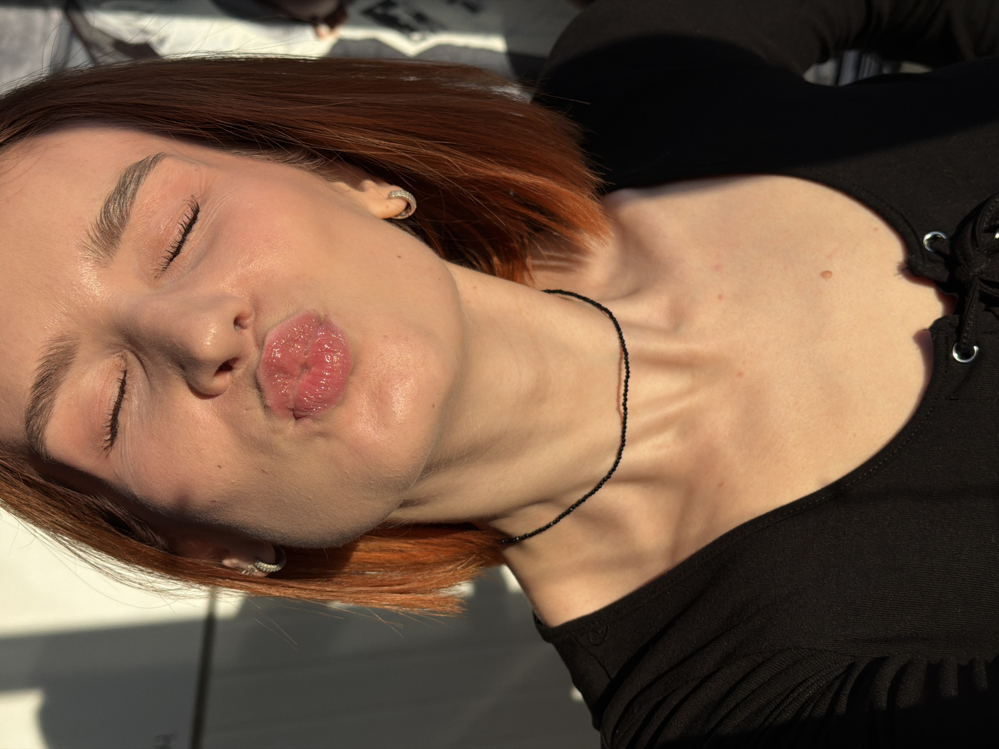 A young woman with red hair and closed eyes puckers her lips in a kiss. She wears a black shirt and a delicate black necklace. Silver hoop earrings are visible in her ears. The light highlights her skin, casting shadows across her face and upper body. The location, Chaikovsky, Russia, cannot be determined from the image alone.