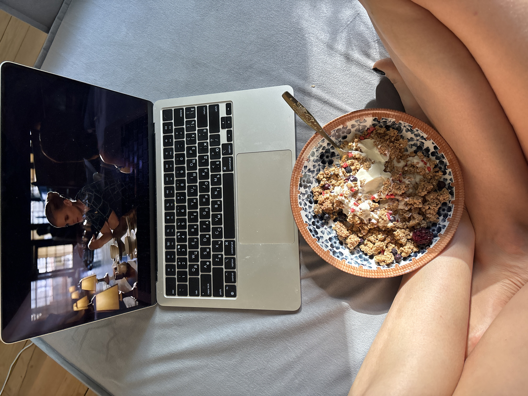 A top-down, medium-shot depicts a woman with crossed legs in a casual setting, seemingly on a bed, with a bowl of cereal placed in her lap. A laptop sits next to her, screen open, displaying an image that is difficult to discern, though it appears to show a person in an interior setting with lighting fixtures. The laptop features a keyboard with Cyrillic characters, indicating a Russian or Eastern European origin. The bowl contains granola, berries, and a dairy-like substance, presumably yogurt or milk, with a spoon resting inside. The setting is a light-grey bed. There is no specific indication that the location is Chaikovsky, Russia, nor any information to suggest the image was taken there.