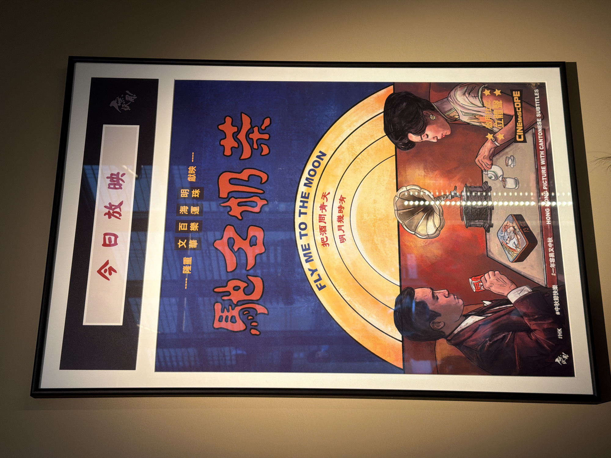 A framed poster hangs on a beige wall. The poster's design is a collage-style layout, incorporating imagery reminiscent of old Hong Kong cinema. On the left side of the poster are vertical rows of Chinese characters in red and black with white borders. The right side features illustrations of a man and a woman seated at a table with a vintage gramophone. A half-circle of gradient orange and yellow separates the two sides. The title "FLY ME TO THE MOON" is inscribed on the orange portion. The bottom right corner contains the text "HONG KONG PICTURE WITH CANTONESE SUBTITLES." The poster is encased in a black frame with a white mat. No location or people are visible beyond what is shown in the poster.