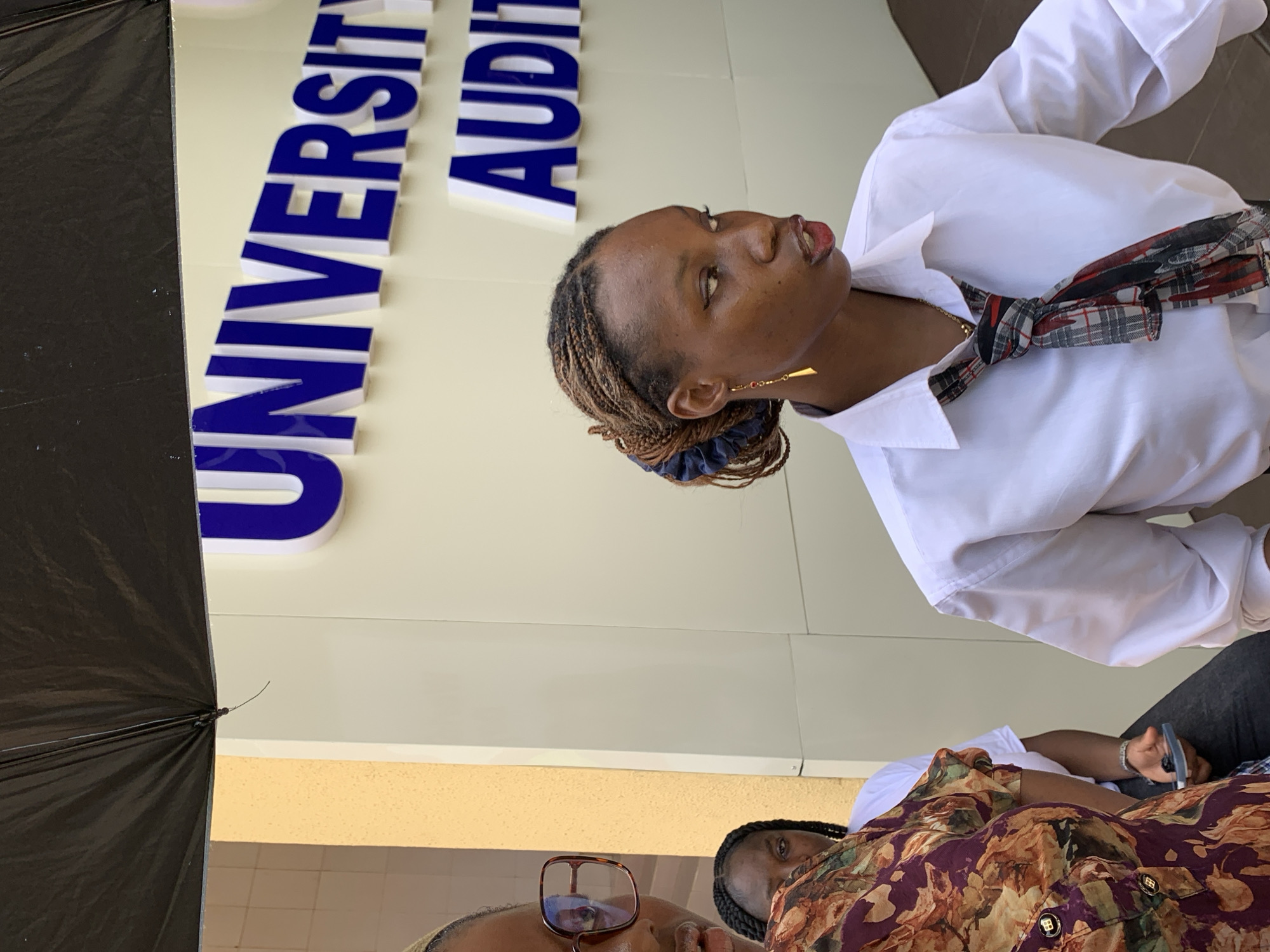 The image captures a close-up of three individuals outdoors, under bright daylight, with a building facade in the background. The main subject is a young Black woman positioned in the mid-ground, wearing a white collared shirt and a plaid scarf tied around her neck. She has braided hair, pulled back with a blue scrunchie, and wears a long, gold-colored earring on her visible ear. Her head is tilted slightly upwards, and she appears to be looking towards the right side of the frame with a focused expression.
In the foreground, partially visible on the left, is a black umbrella. Also in the foreground, another person's head and shoulders are visible, wearing glasses and a floral-patterned top in shades of maroon, orange, and green. A third person is partially visible behind the main subject, wearing dark clothing and holding a pen, suggesting they might be writing or filling out something.
The background features a light-colored building with large, raised blue letters spelling out "UNIVERSITY AUDIT," which likely indicates "UNIVERSITY AUDITORIUM." The setting is outdoors, likely a public or campus area in Ilorin, Nigeria, given the provided context. The bright lighting suggests it is daytime. The people appear to be either waiting, observing an event, or engaged in some form of registration or administrative process.