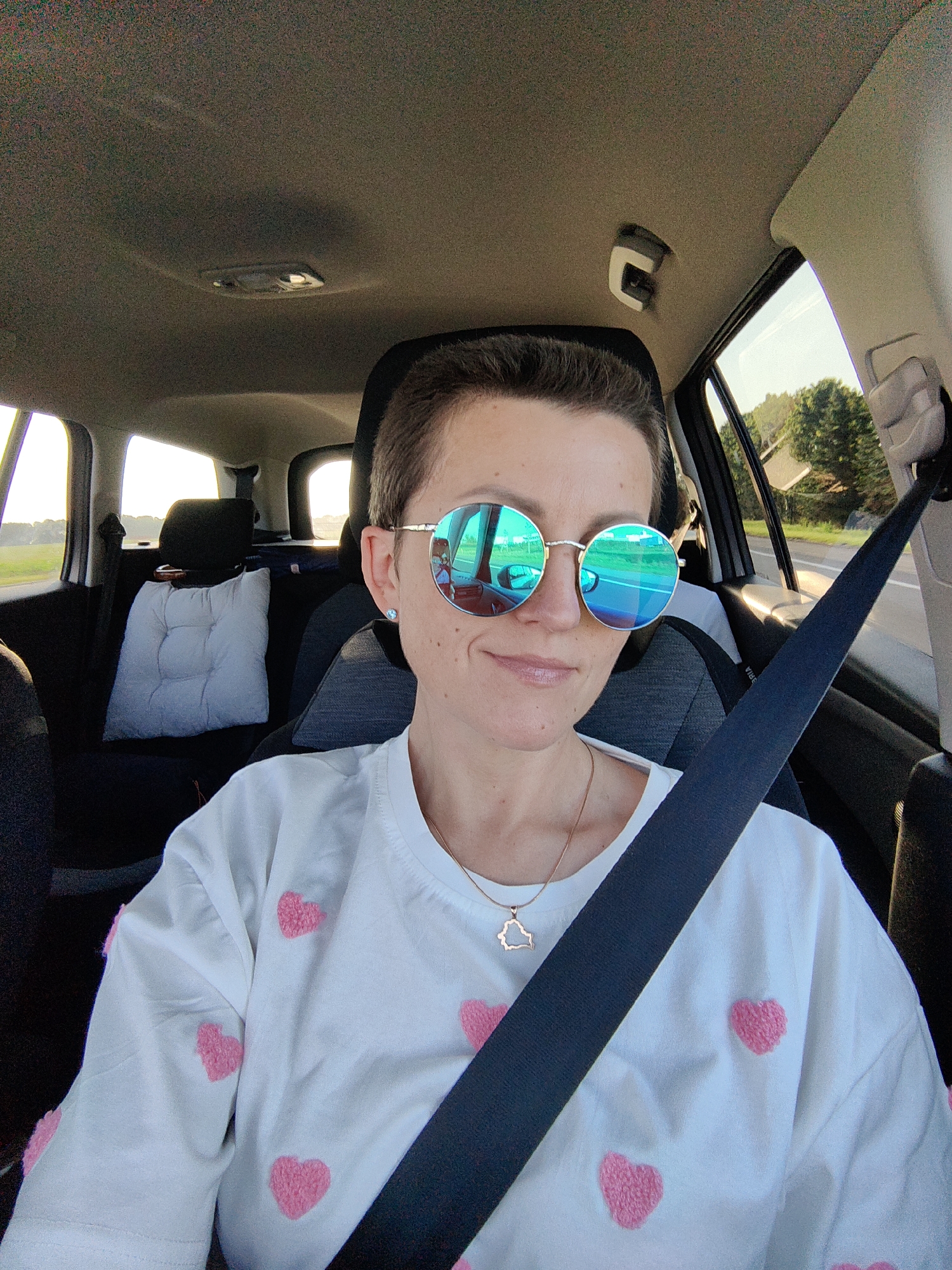 A woman with short, dark hair and round, mirrored sunglasses is in the passenger seat of a car. She is wearing a white t-shirt with pink heart patterns and a silver necklace with a pendant shaped like the country of Belarus. The car's interior is visible, including the headrest of the driver's seat, a white cushion on the back seat, and the car's ceiling with a light fixture. The background shows trees and a road through the car windows, suggesting an outdoor setting, possibly in a rural area. The scene appears to be a candid photograph taken during daylight hours. Given the presence of the Belarus-shaped pendant and the request for a location within Міханавіцкі сельскі Савет, Belarus, it is plausible the woman is traveling within or near this region.