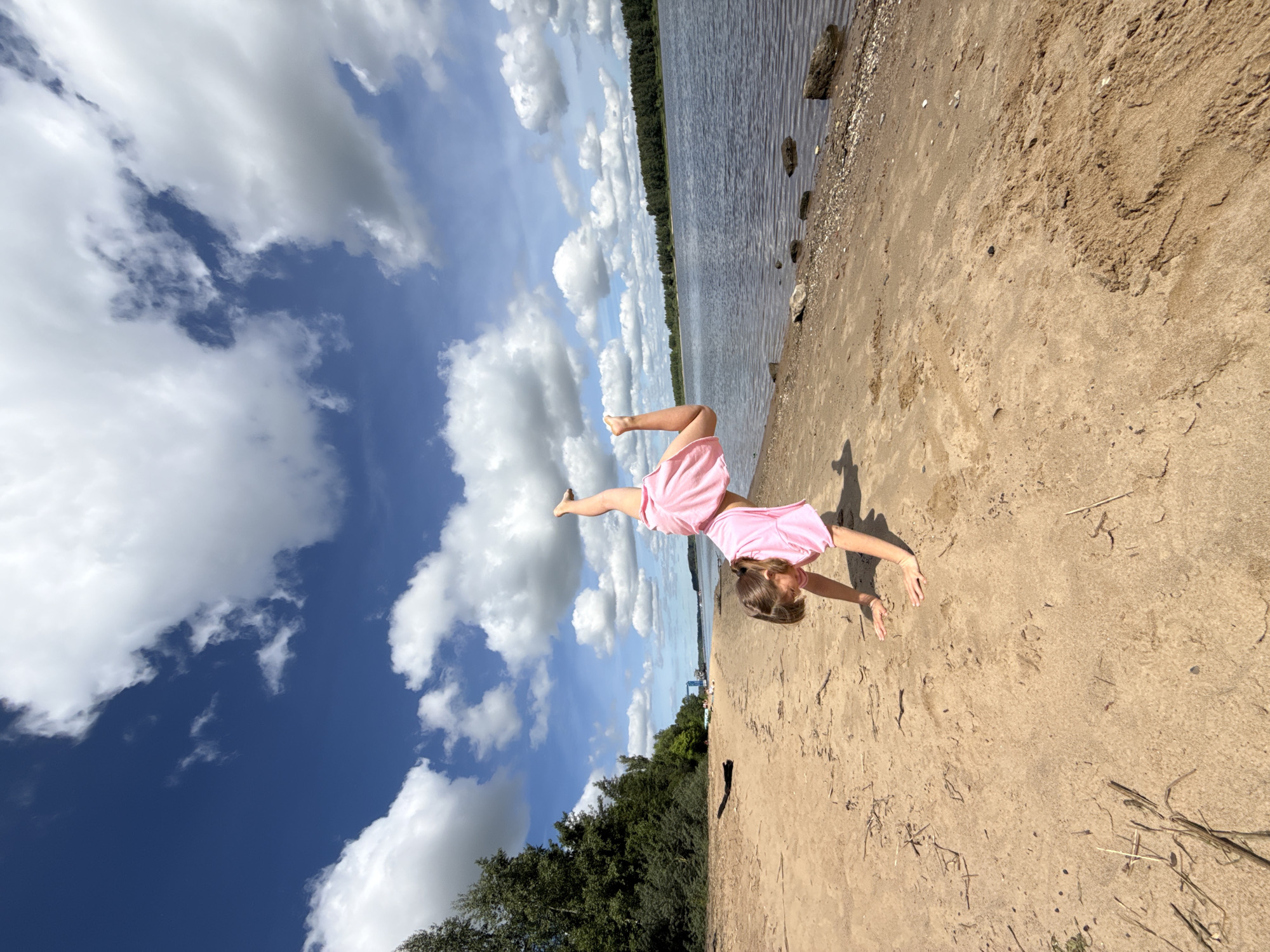 The image captures a vibrant outdoor scene on a sunny day at a sandy beach in Rusanovo, Russia. A young girl, dressed in a pink t-shirt and pink shorts, is depicted in dynamic motion, seemingly performing a cartwheel or an acrobatic pose. Her hands are placed on the light brown sand, and one leg is extended high into the air, showcasing her flexibility and active play. Her head is angled downwards, partially obscuring her face as she focuses on her movement.
The setting features a wide expanse of light brown sand, with visible textures and a few scattered pieces of dried vegetation. To the right of the girl, a calm body of water stretches into the distance, reflecting the bright sky. The opposite shore is lined with a dense forest of green trees, suggesting a natural and serene environment. Small rocks and pebbles are visible along the water's edge on the sandy bank.
Above, the sky is a striking deep blue, adorned with numerous fluffy white cumulus clouds, indicating clear and pleasant weather conditions. The bright daylight illuminates the scene, enhancing the cheerful and energetic atmosphere. The image conveys a sense of playful freedom and joy, capturing a moment of active outdoor recreation. No visible text is present in the image.