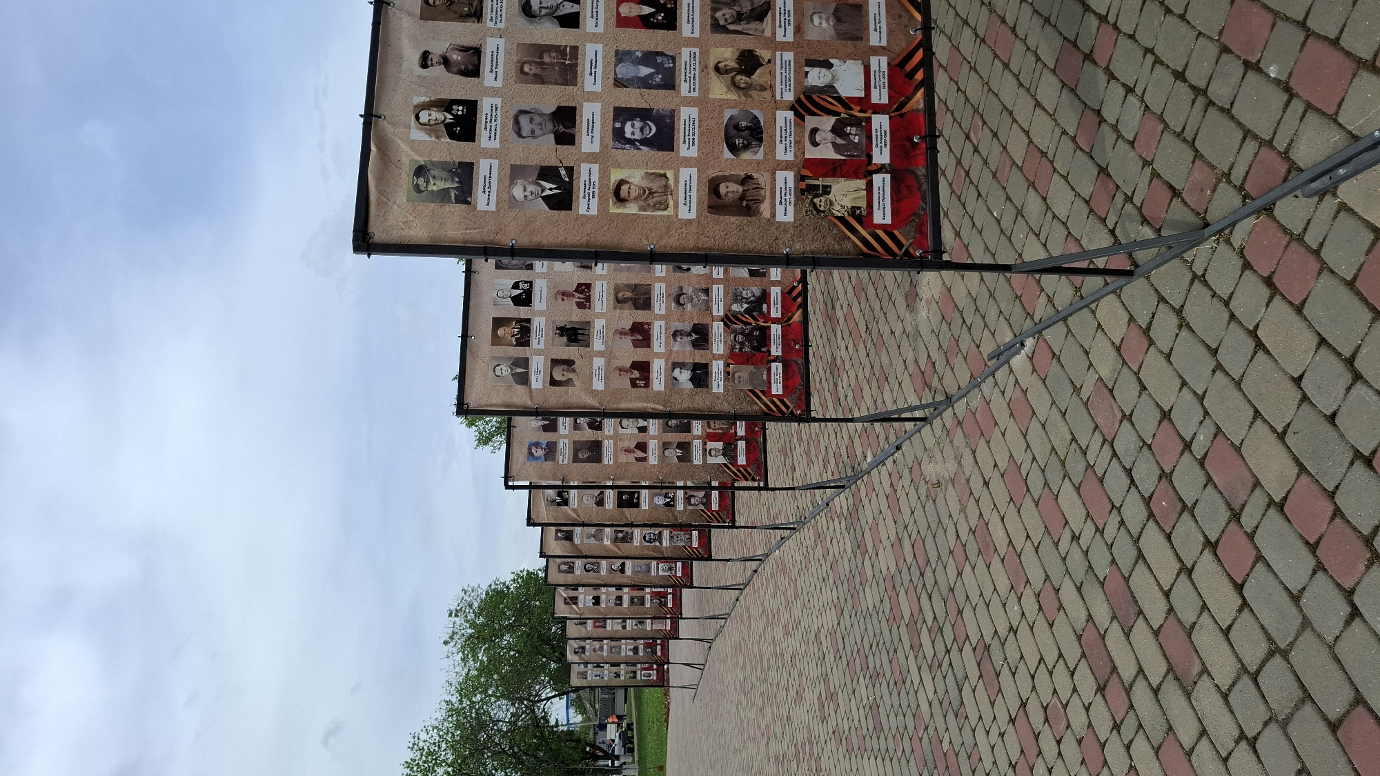 The image shows a series of memorial banners lined up along a brick walkway. Each banner displays multiple black and white portraits with names and dates. The banners are supported by dark metal stands. The walkway is paved with alternating rows of grey and red-toned bricks. A tree with green foliage is visible to the left of the banners. The sky overhead is cloudy. The location is a city of Unknown, Unknown.