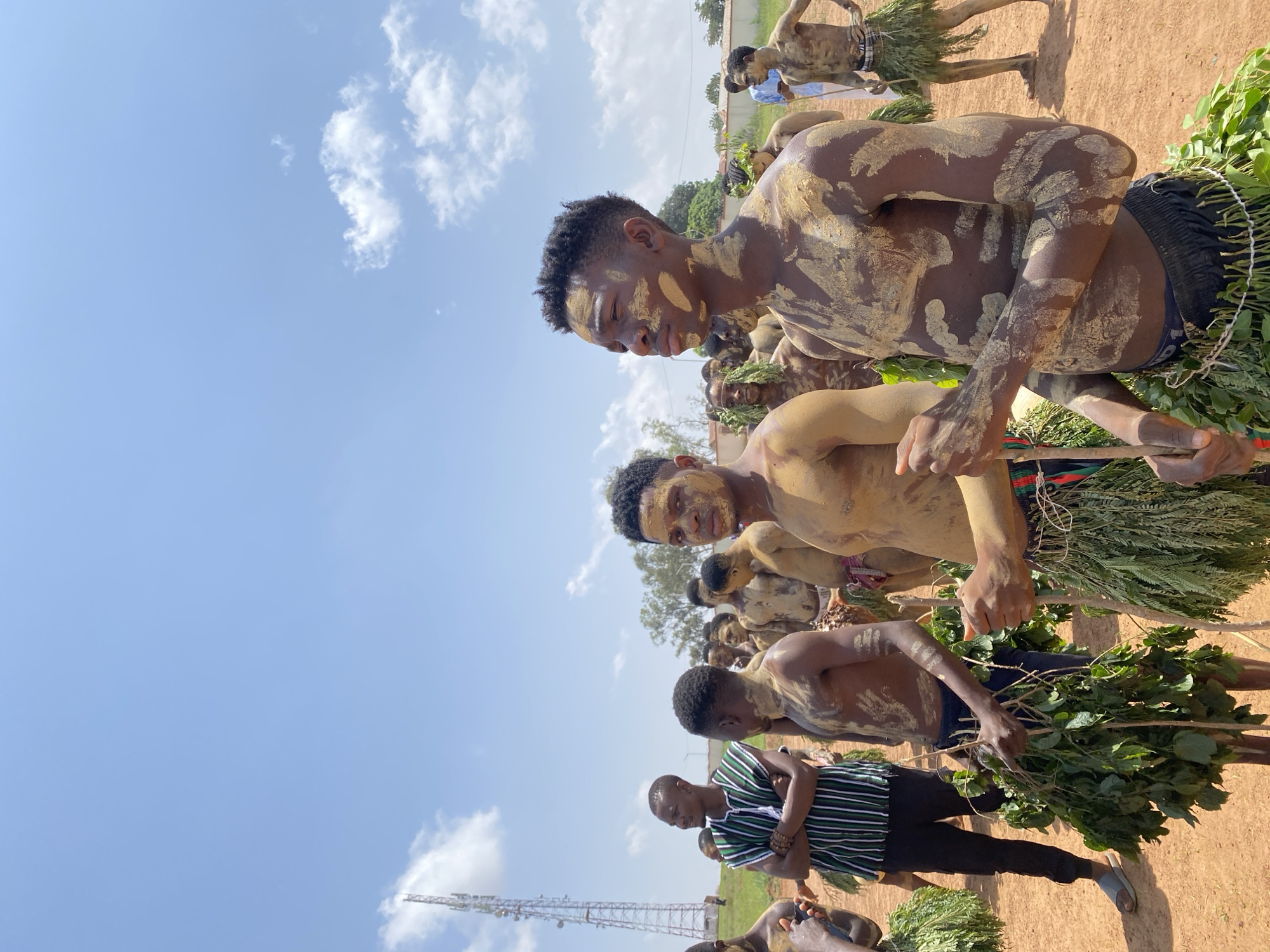 A group of bare-chested men are outdoors on a dirt ground on what appears to be a sunny day, possibly in Durumi, Nigeria. They are wearing what appears to be mud or clay markings on their skin. Some are carrying branches with green leaves and others have green leaves around their waists. One man in the foreground is wearing a striped shirt with dark pants and sandals. A tall communication tower is visible in the distance under a bright blue sky with a few clouds.