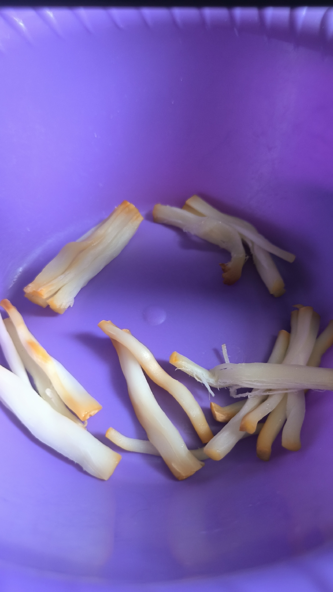 The image shows a close-up view of several white and orange strands of food, likely mushrooms, scattered in a purple bowl. The bowl appears to be plastic with a textured rim. The food items are elongated and appear to be segmented, with some strands fanned out and others clustered together. The lighting suggests an indoor setting, possibly a kitchen. There is no visible text in the image. The overall impression is of ingredients prepared for cooking or serving.