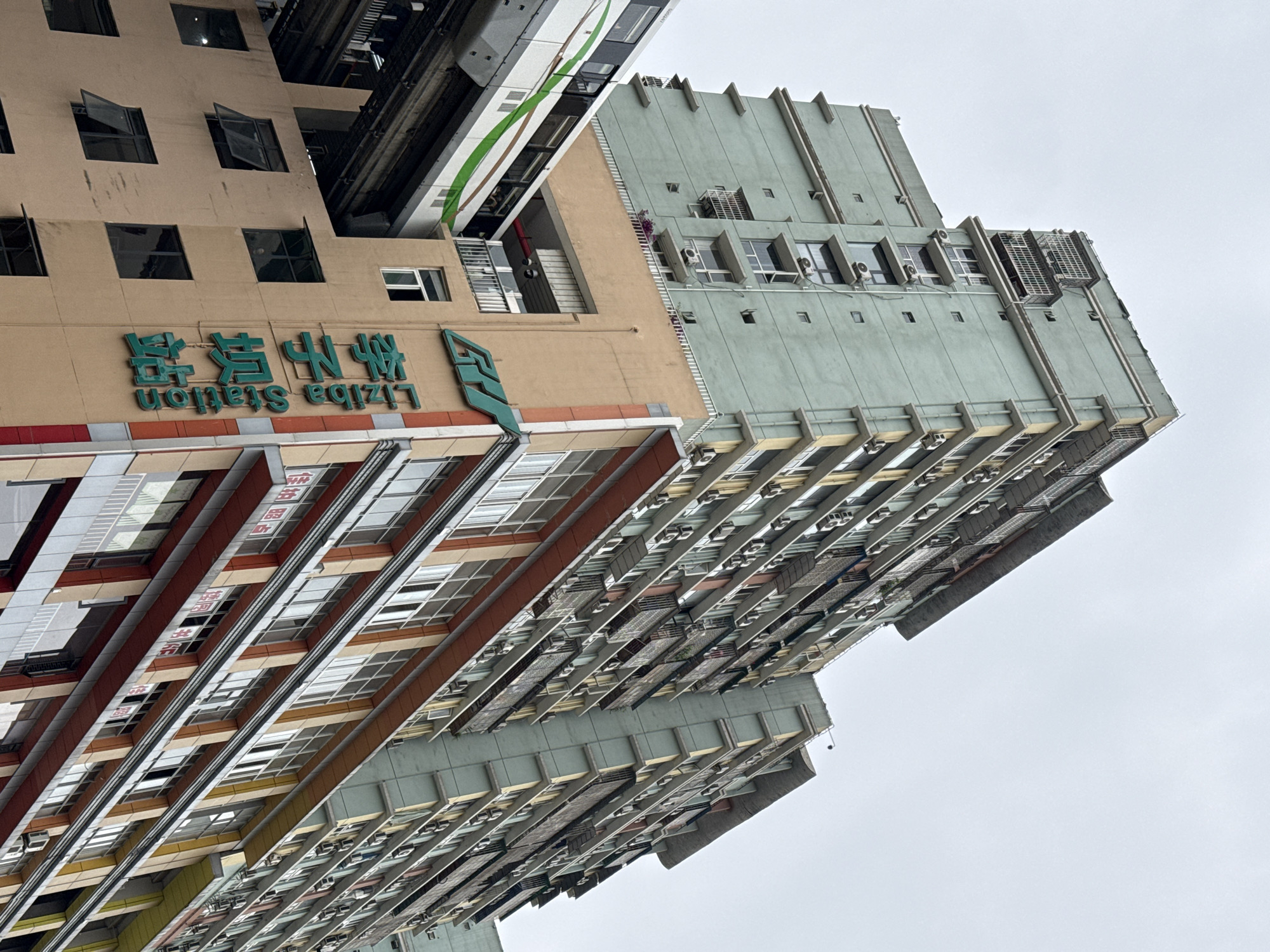 The image captures a dense urban landscape featuring several multi-story buildings and a monorail passing through. The original image is rotated 90 degrees counter-clockwise, showing a vibrant, vertically oriented scene.
In the foreground, a prominent beige building with green accents dominates, featuring multiple square windows, some of which are open. Affixed to its facade is large green signage displaying "Liziba Station" in English and its Chinese characters (李子坝站). The monorail, a sleek train with green and white stripes, is visible on an elevated track, appearing to either pass directly through a section of this beige building or very closely adjacent to it.
To the right of the beige building, another tall structure with a light grey-green facade rises. This building exhibits a more uniform, industrial-like design with numerous smaller windows and many external air conditioning units mounted beneath them. Further to the right and extending into the background, other multi-story buildings with various architectural styles, extensive window arrangements, and numerous air conditioning units can be seen, contributing to a sense of high-density urban living.
The sky is overcast, indicating a cloudy daytime setting. The overall scene depicts a dynamic urban environment, typical of modern cities, with infrastructure like the monorail integrated closely with residential and commercial buildings. The visible text clearly identifies the location as Liziba Station, famous for its unique monorail design in Chongqing, China.