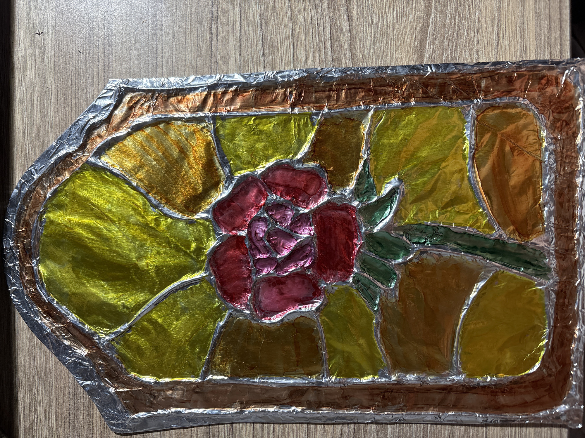 The image displays a homemade stained-glass-style artwork of a rose, created from colorful metallic foil. The artwork is framed with what appears to be brown and silver foil, mimicking leaded glass. The rose is depicted in shades of red and pink, with green foil for the stem and leaves. The background is a patchwork of yellow and orange foil, suggesting petals or light. The entire piece is laid on a wooden surface, likely a table, with a visible wood grain pattern. The lighting casts shadows, highlighting the textured, crinkled surface of the foil. There are no people or animals present in the image. The artwork is positioned on the wooden surface, and no specific location within Panagyurishte, Bulgaria, is indicated or inferable from the image itself.