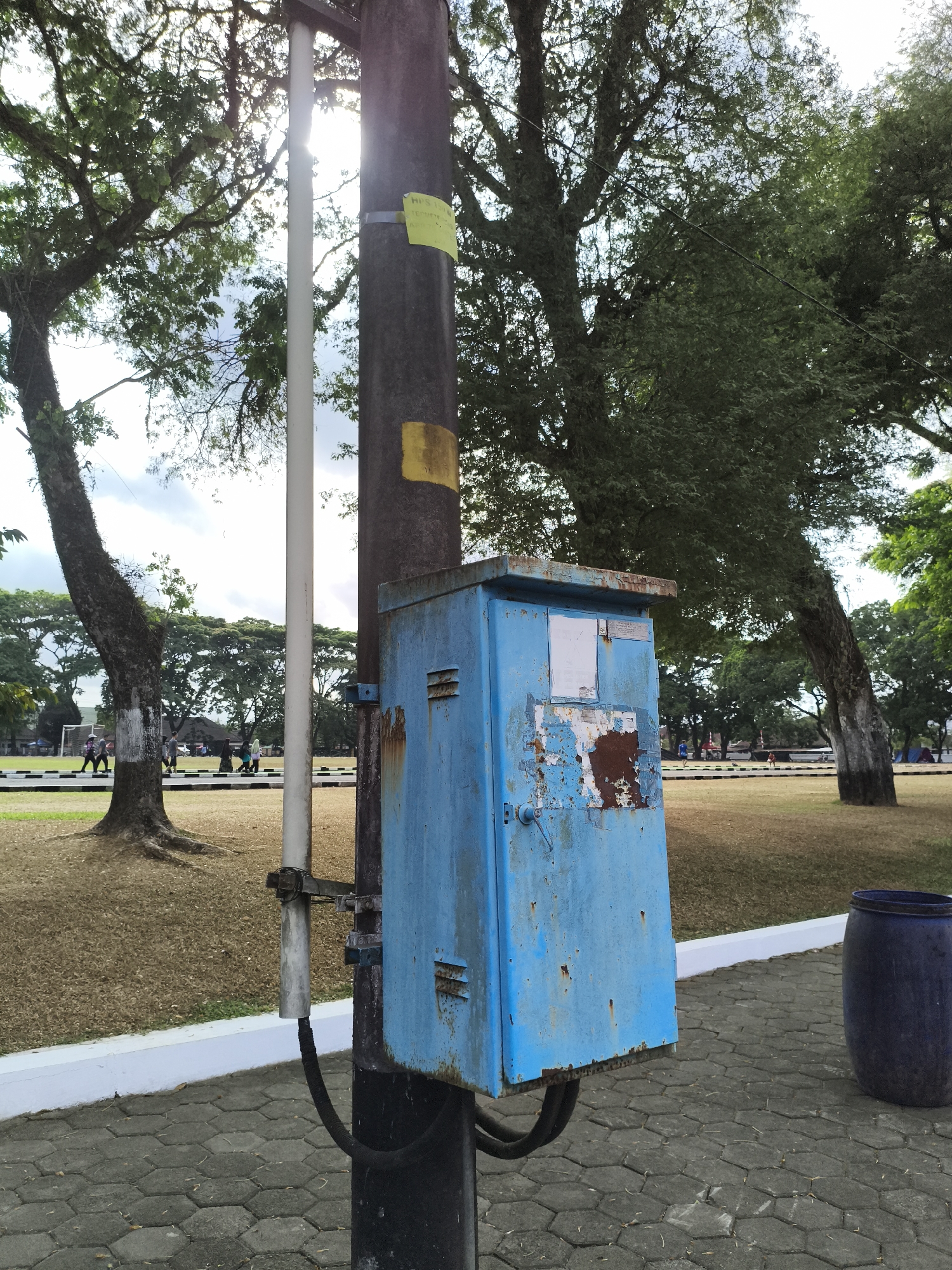 A weathered, blue metal electrical box is attached to a dark, cylindrical utility pole in what appears to be a park or open grassy area. The electrical box, showing signs of rust and peeling paint, has a handle and what seem to be ventilation slits. A yellow sticker and a yellow rectangular marking are present on the pole above the box. A white pipe runs vertically alongside the pole, and black conduits emerge from the base of the box, feeding into the ground. The ground in the foreground is paved with grey hexagonal interlocking blocks.
In the background, a wide expanse of dry, light brown grass is visible, bordered by a white curb. Beyond the grassy area, several large trees with dense green foliage stand, some with thick, gnarled trunks. Sunlight streams through the trees, creating dappled light and glare, particularly in the upper left quadrant of the image. In the far distance, a few individuals can be seen walking and possibly engaging in recreational activities on a lighter colored surface, suggestive of a sports field. To the right of the scene, a large, dark blue plastic barrel is positioned on the paved ground. The overall environment suggests an outdoor public space. The location is identified as Taman Kyai Langgeng in Magelang, Indonesia.