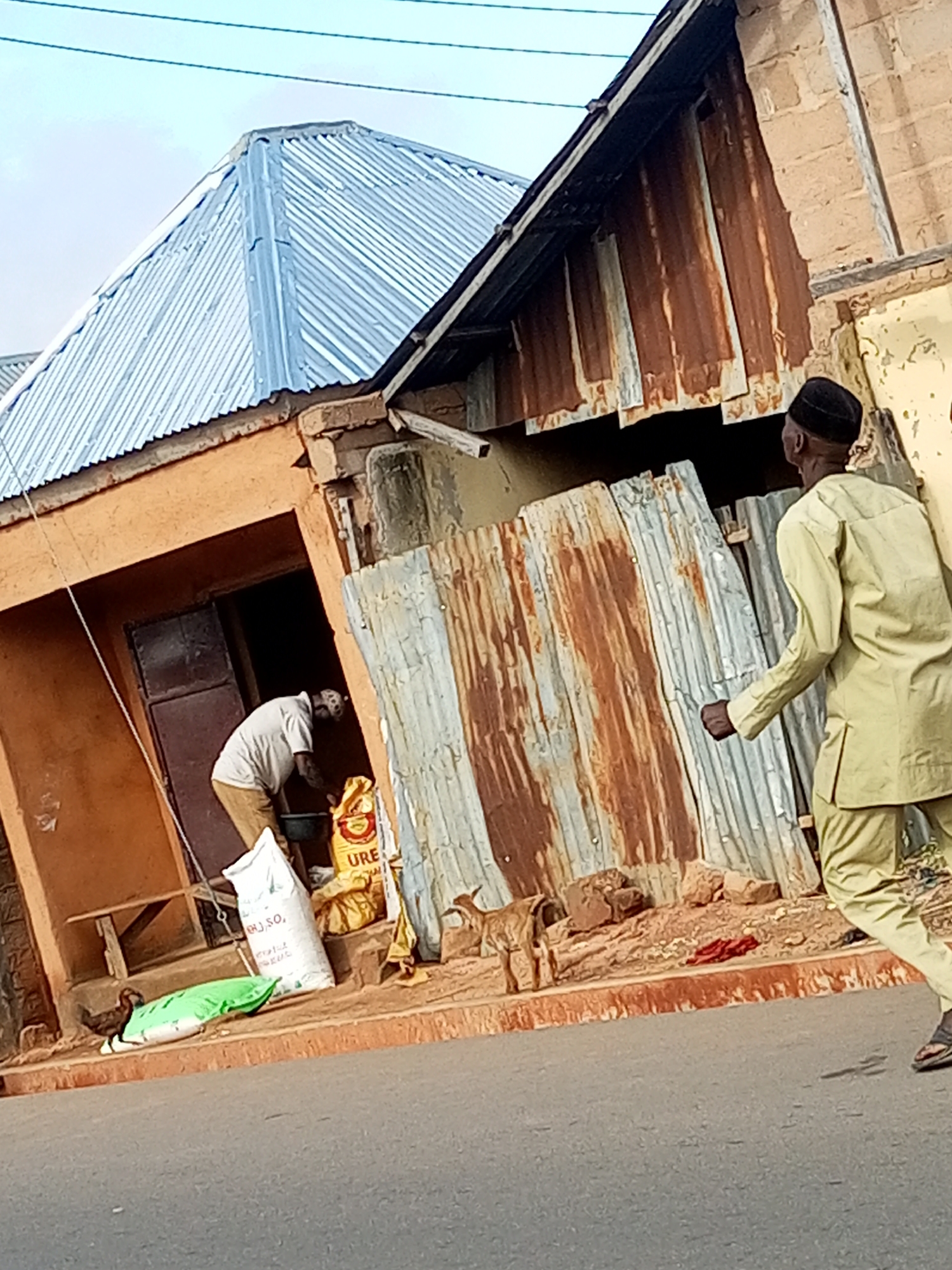 The image depicts a scene on a street in Gombe, Nigeria, with a focus on a building that appears to be a shop or dwelling. The building has a corrugated metal roof, partially rusted, and walls made of mud brick and corrugated metal sheeting. To the left, a man in a light-colored shirt and brown pants is visible inside an opening, possibly a doorway, attending to a large yellow bag, possibly containing fertilizer, and other items on the ground. A green bag is also on the ground near him. A small brown goat stands near the building's entrance. To the right, a man in a pale green traditional outfit, possibly a kaftan, is walking away from the building, facing away from the camera and towards the right of the frame. The street in the foreground is paved asphalt. The sky is overcast. The overall impression is one of everyday life in a somewhat unkempt urban environment.