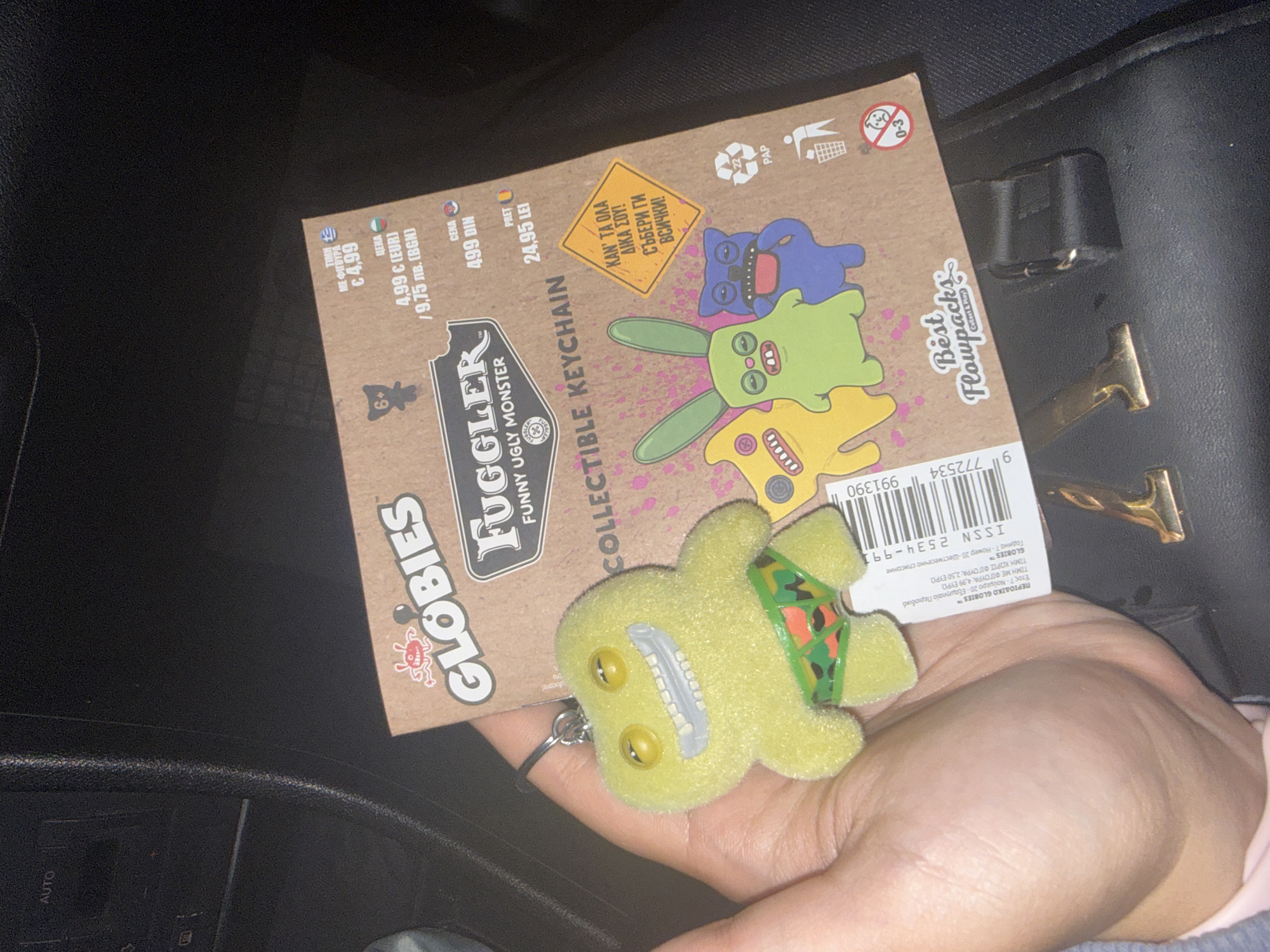 The image shows a hand holding a Globies "Fuggler" collectible keychain. The keychain is yellow and depicts a monster with a green and orange patterned diaper. The keychain is attached to a metal keyring. The packaging for the keychain is brown cardboard. The packaging states "Globies Funny Ugly Monster Collectible Keychain". The packaging also includes pricing in Euro (€4.99), Bulgarian Lev (9.75 лв.), Serbian Dinar (499 DIN) and Romanian Leu (24.95 LEI). The packaging has a warning label for children aged 0-3. The background includes part of a car interior, and a black leather purse. The location cannot be determined as Okop, Bulgaria from the image.