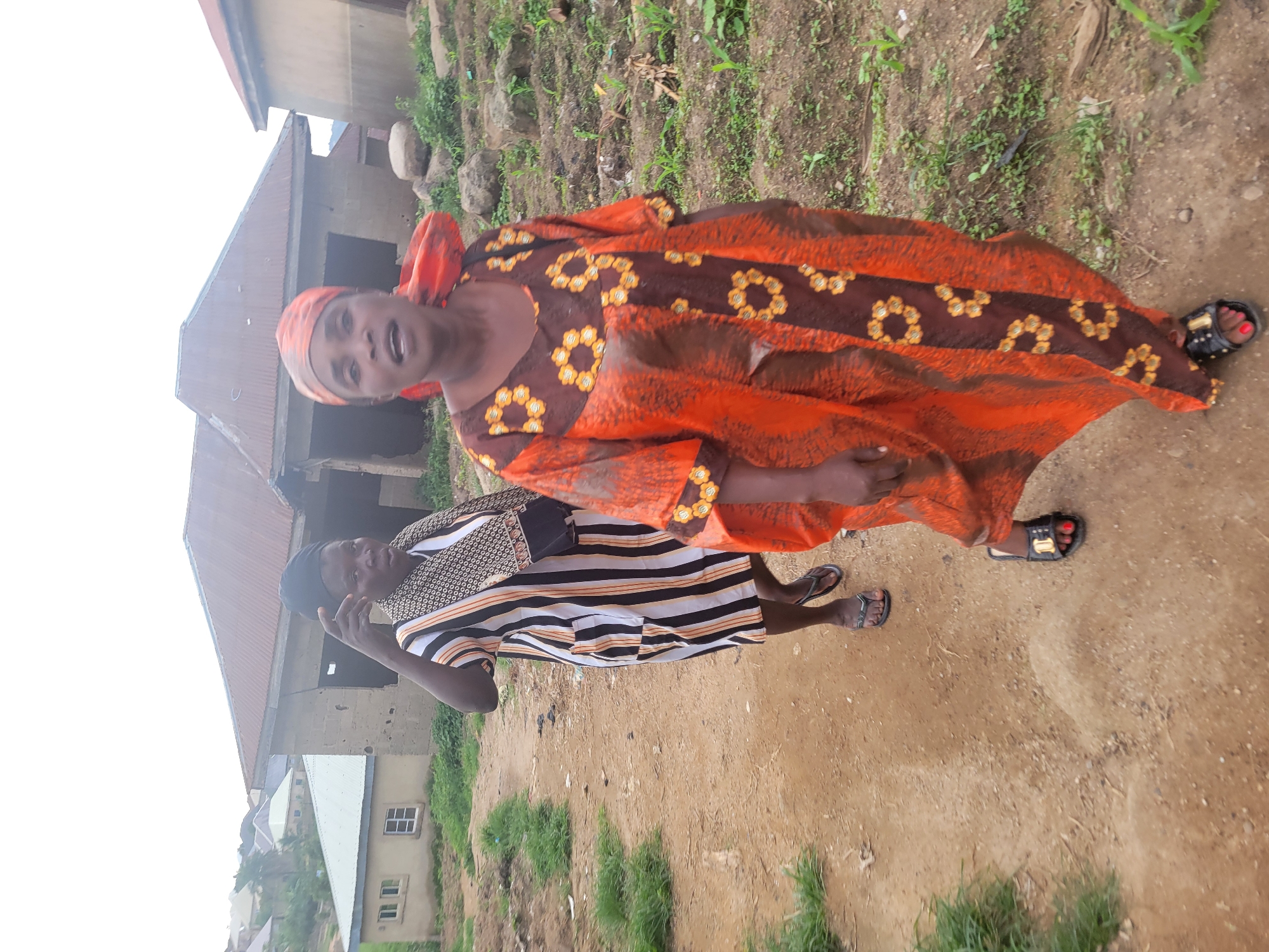 In Madalla, Nigeria, two women walk on a dirt path. The woman in the foreground wears an orange and brown patterned dress and a headscarf. The woman behind her is dressed in a striped tunic and wears a head covering. The background shows several buildings with corrugated metal roofs, some appearing unfinished, and vegetation. The scene depicts a residential area with a mix of constructed and developing structures.